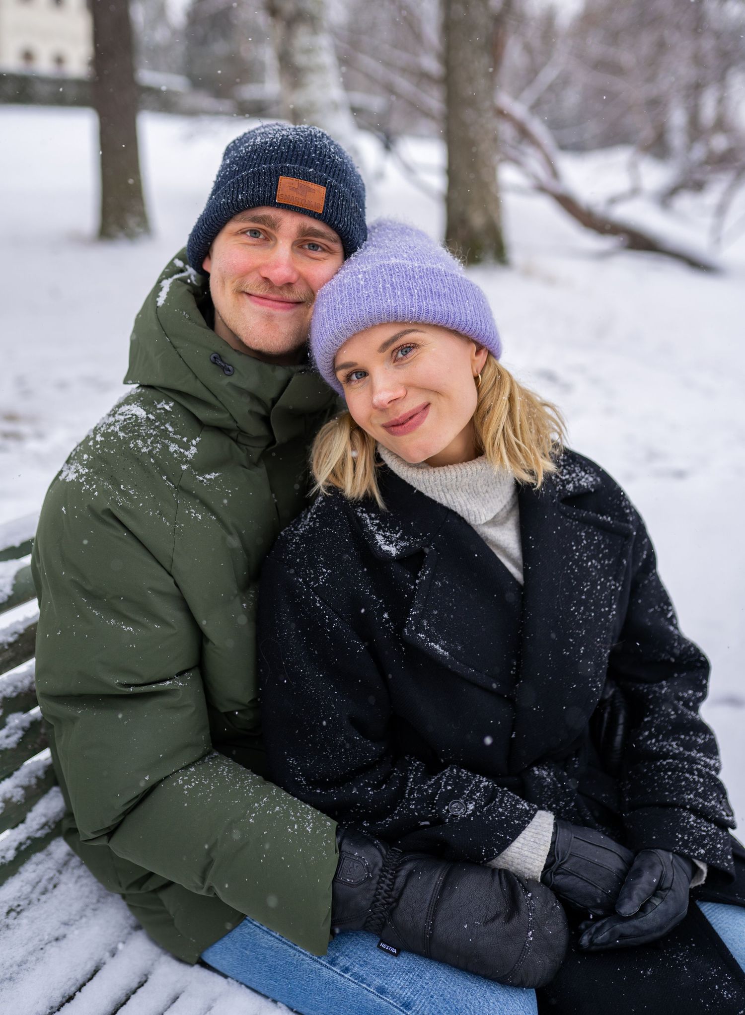 A smiling couple in winter hats and jackets sitting on a snow-covered bench in a park during snowfall.