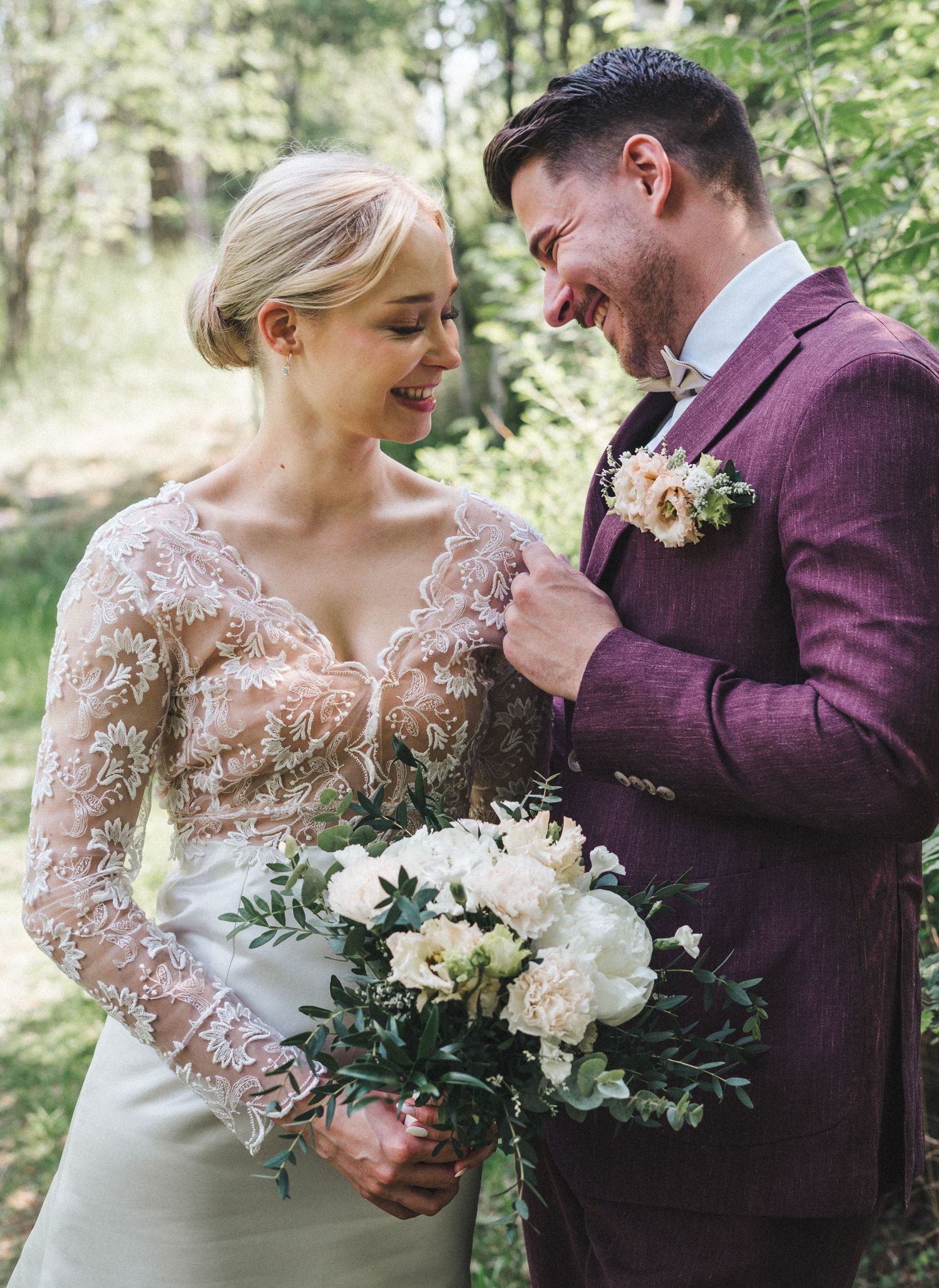 Laughing bride holding white peony bouquet stands close to smiling groom in burgundy suit outdoors.