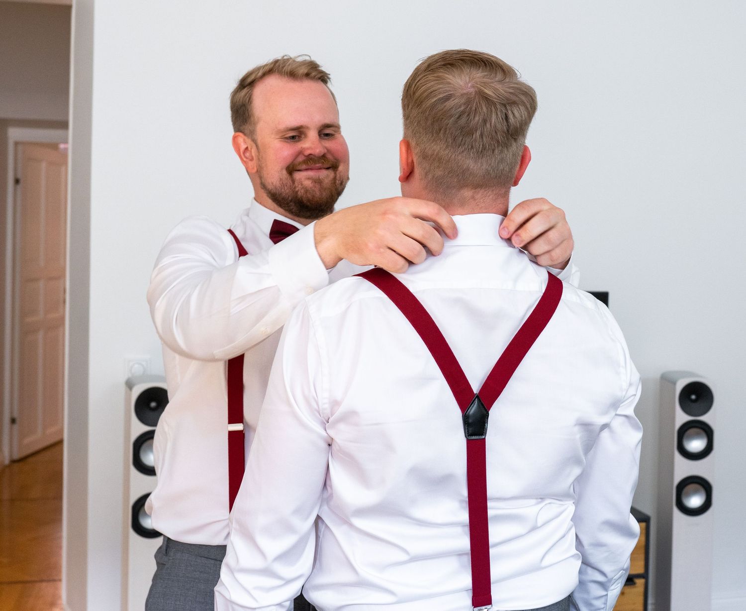 Groomsman in white shirt with burgundy suspenders helping another groomsman adjust his collar before wedding.