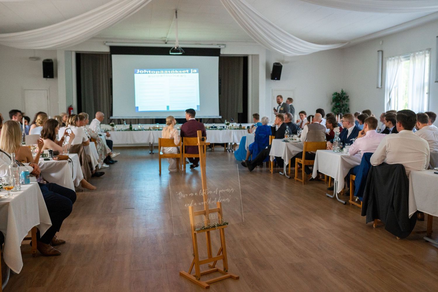 Elegant wedding reception hall with draped ceiling, guests at tables, and presentation screen.