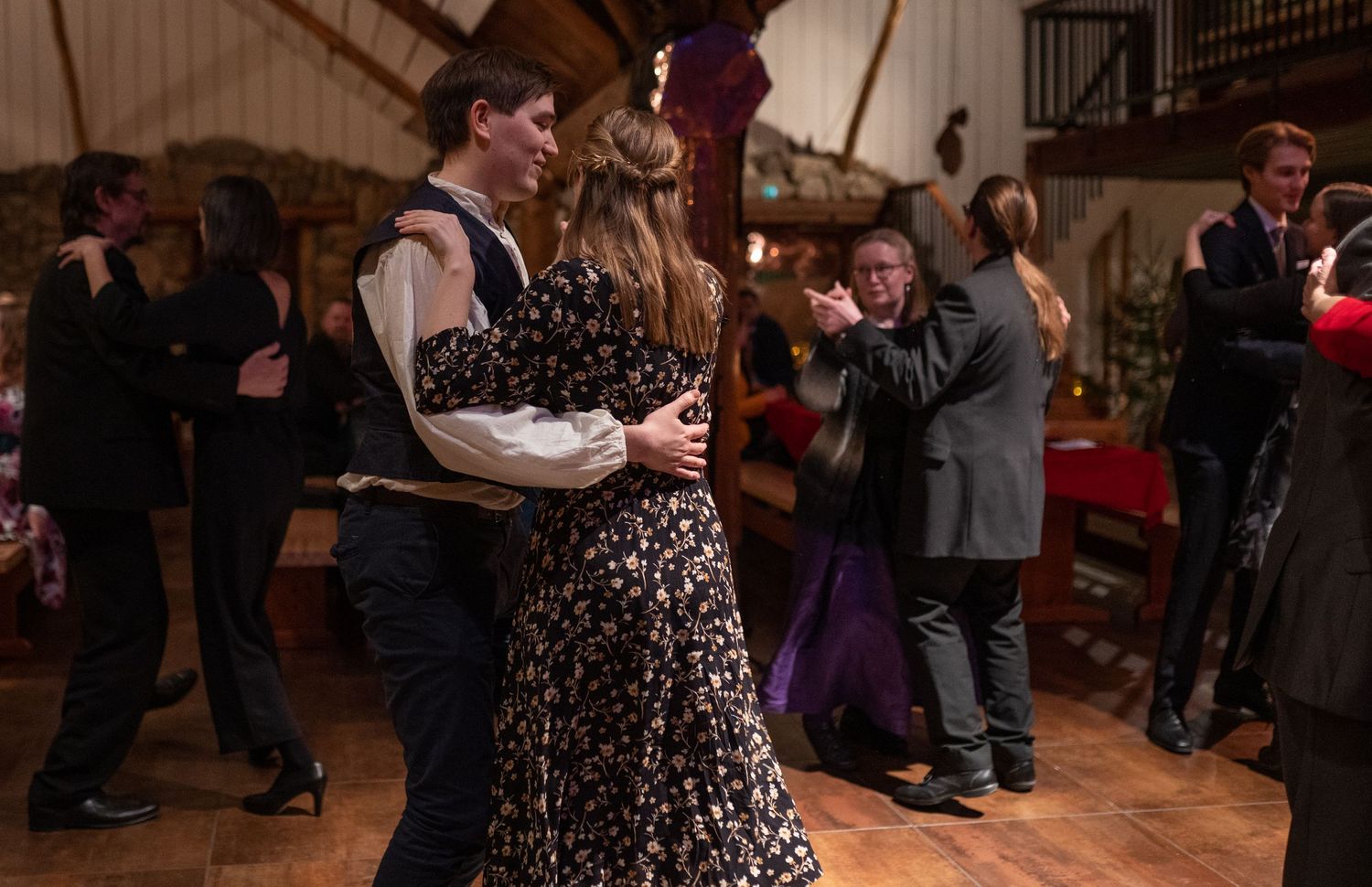 Couples dancing elegantly in formal attire at an indoor evening dance event in a warmly lit hall.