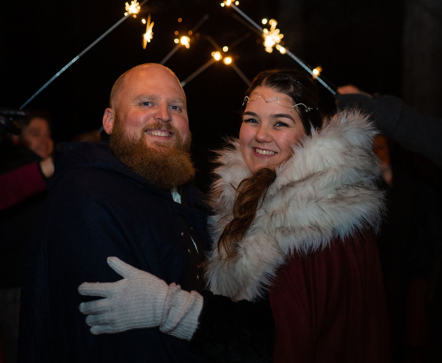 A bearded man and smiling woman pose together outdoors at a festive night event with sparklers glowing in the background.