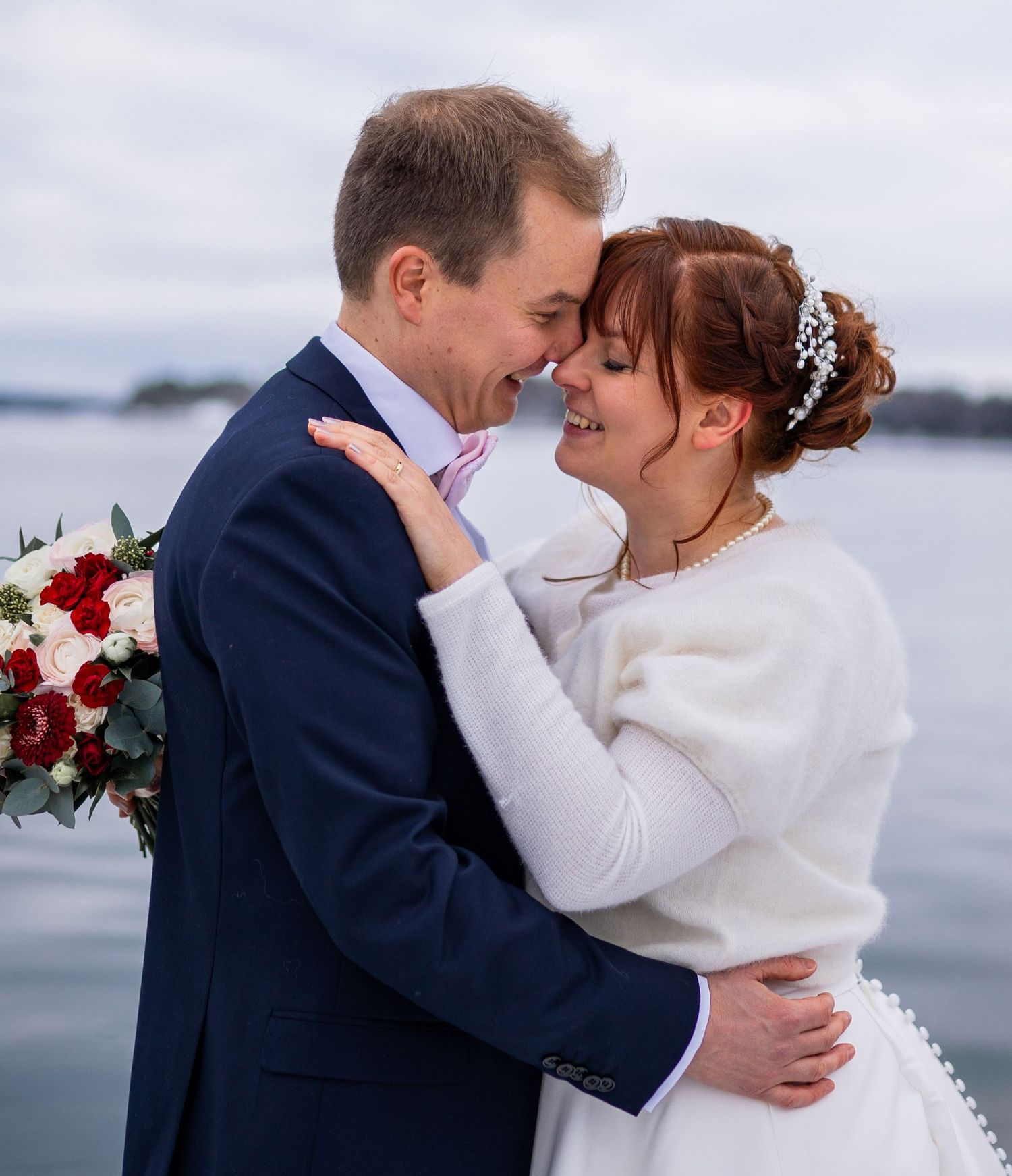 Romantic newlyweds touch foreheads and embrace near a tranquil waterfront on their wedding day.