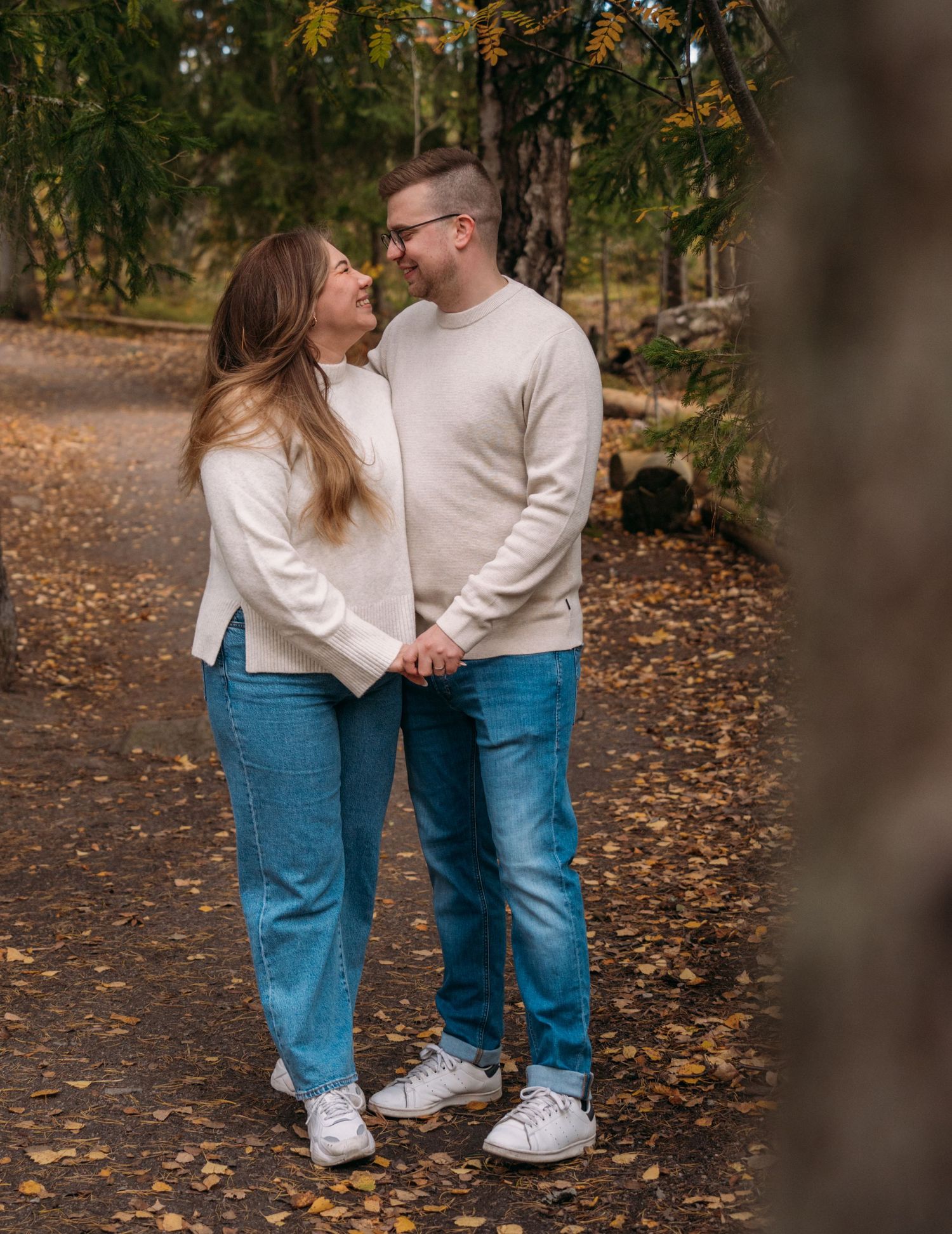 Couple holding hands and smiling at each other in autumn forest, both wearing cream sweaters and blue jeans.