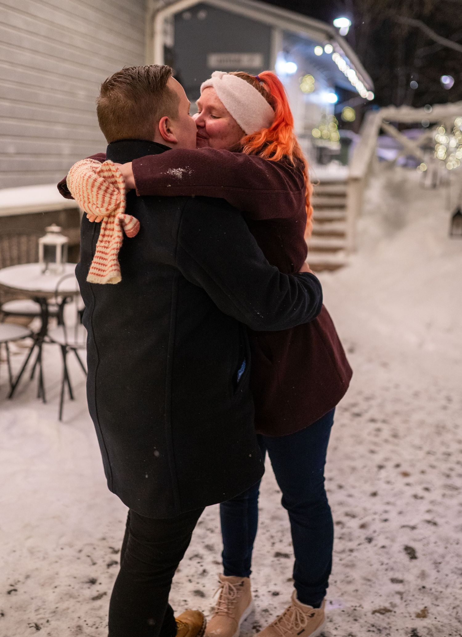 Couple sharing a romantic kiss outdoors in winter snow, woman with orange hair wearing burgundy jacket and pink scarf.