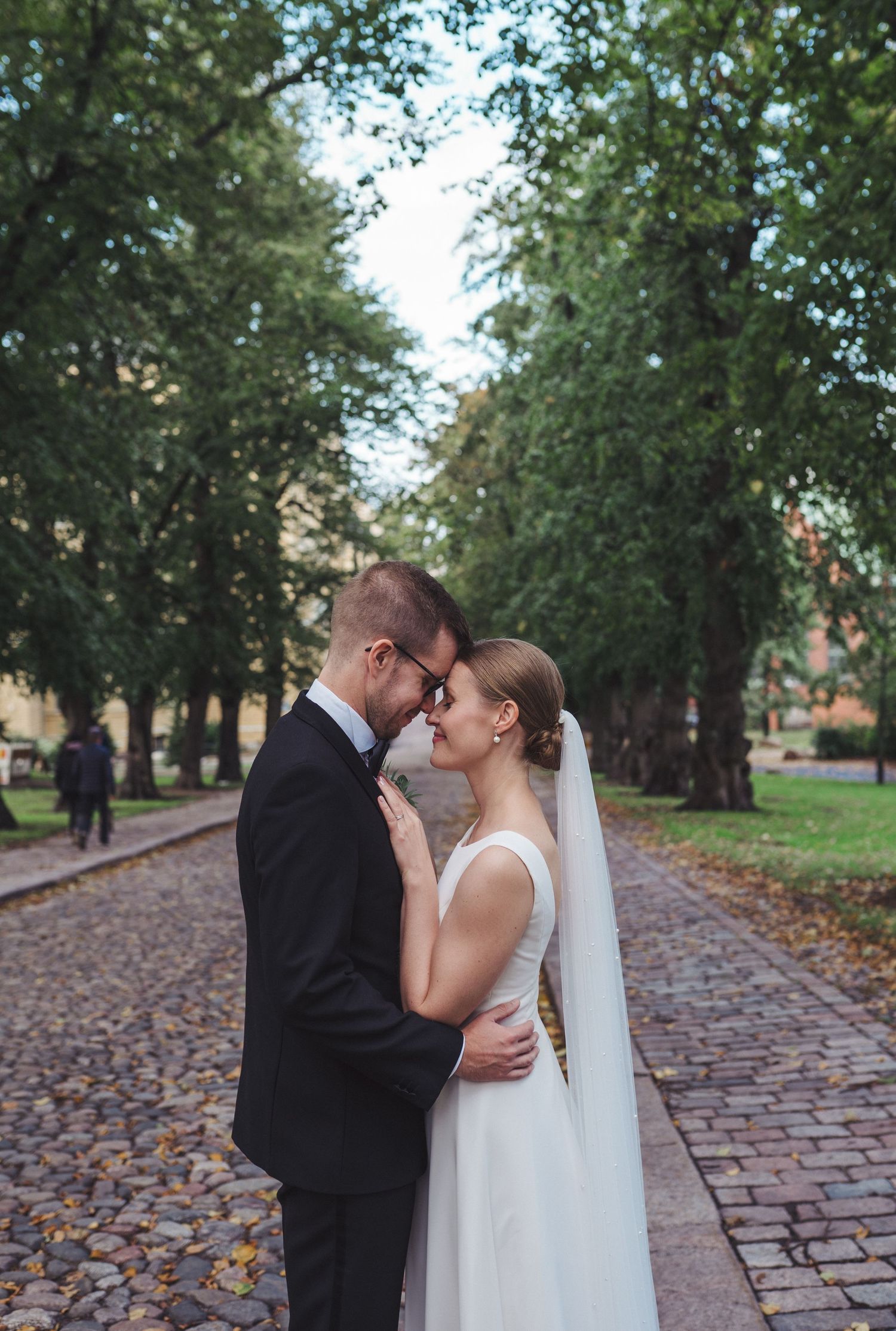 Bride and groom touch foreheads tenderly on a cobblestone tree-lined avenue.