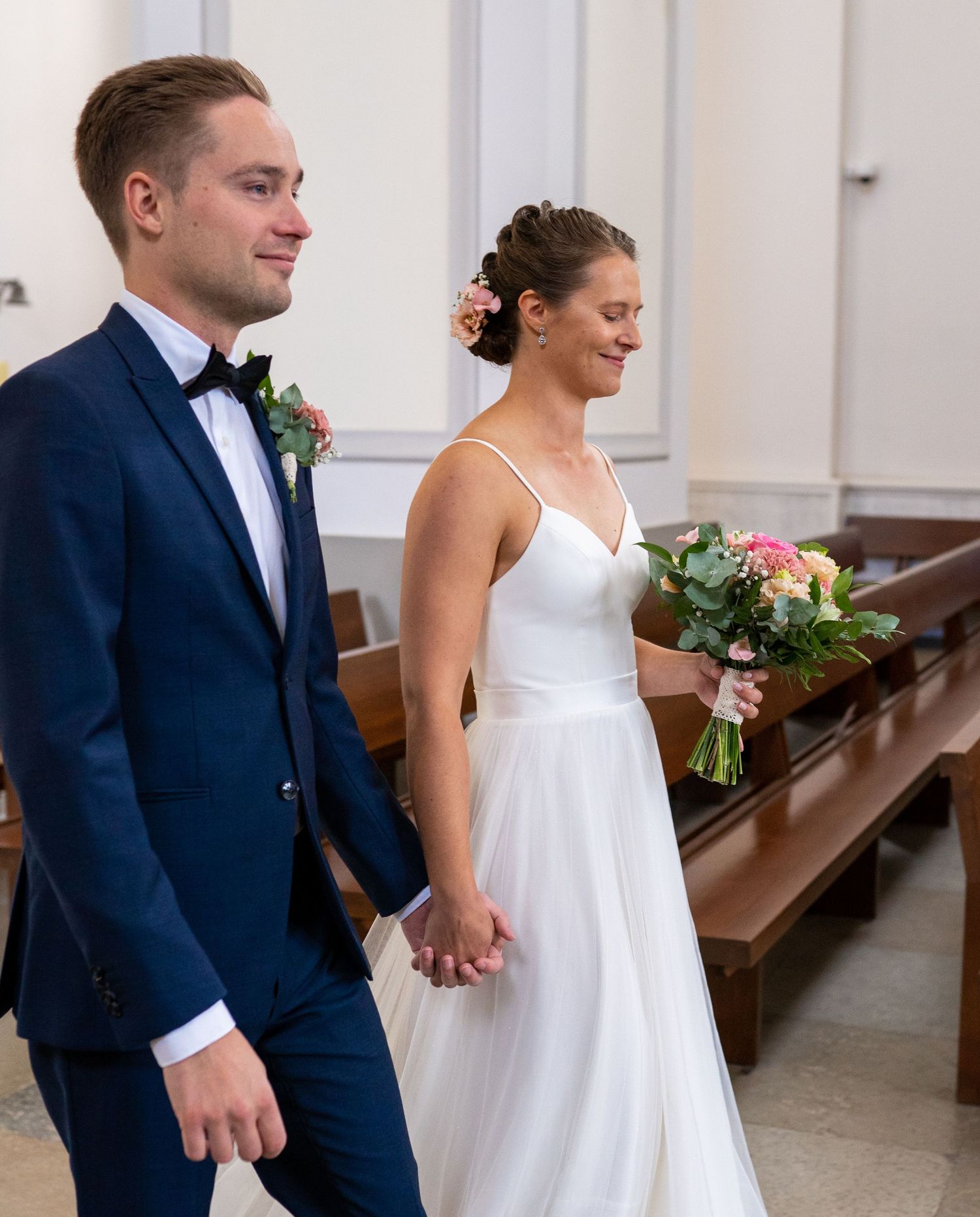 Smiling bride holding a colorful bouquet walking beside groom in navy suit in church.