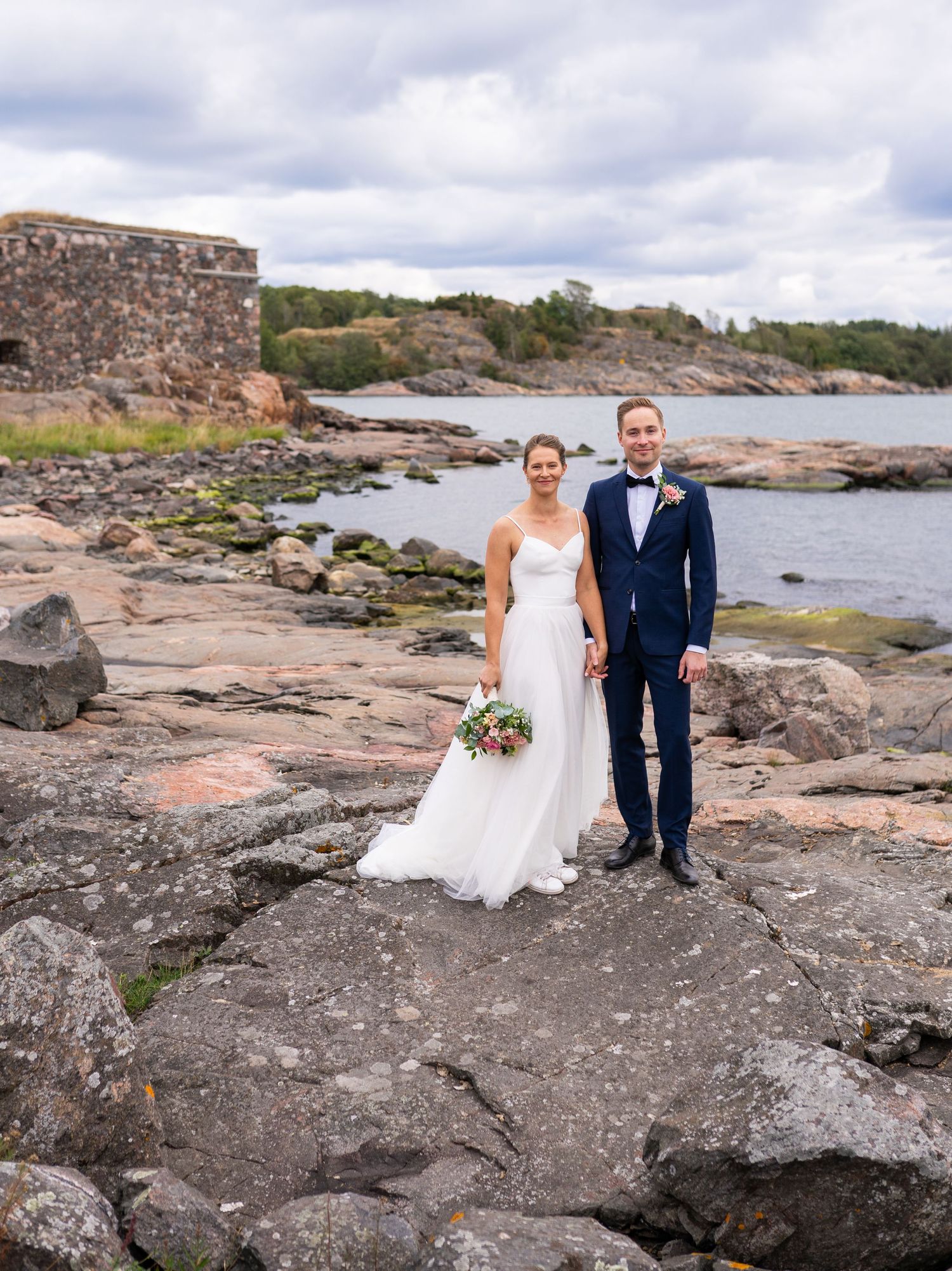 Bride and groom holding hands on rocky coastal terrain, historic stone ruins and sea visible behind them.
