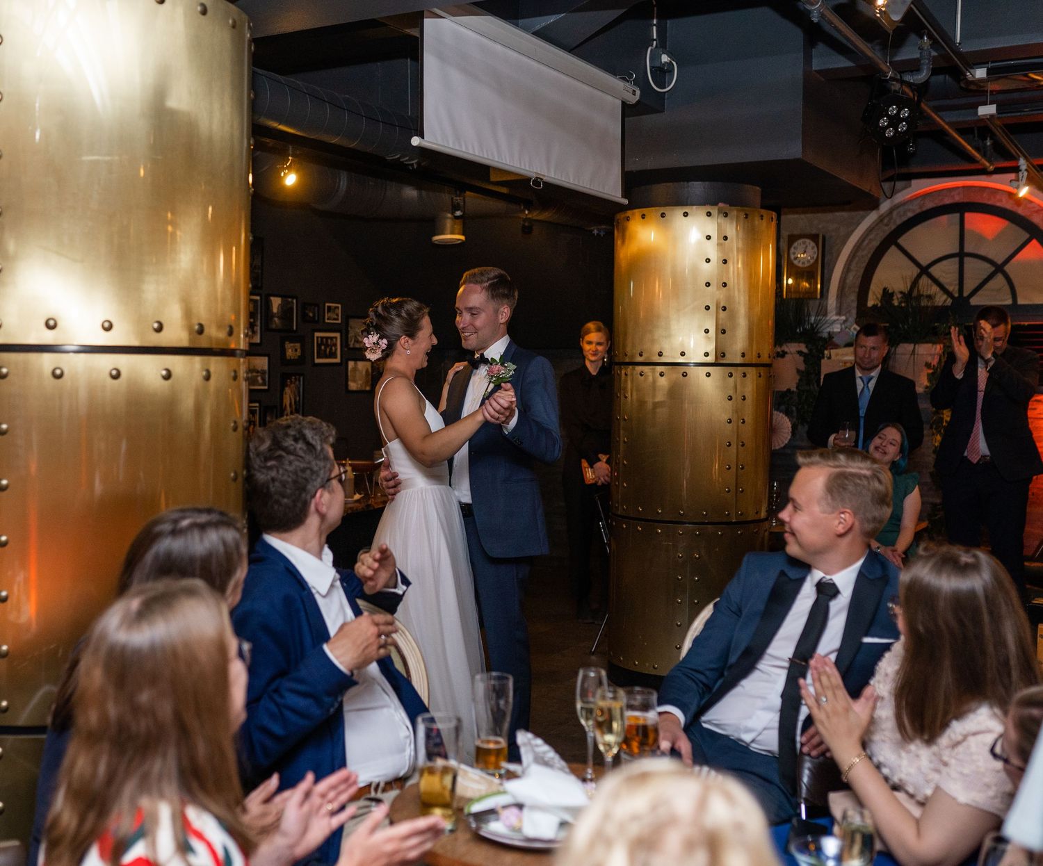 Wedding couple performs first dance as guests clap and celebrate in a sophisticated industrial-style reception venue.