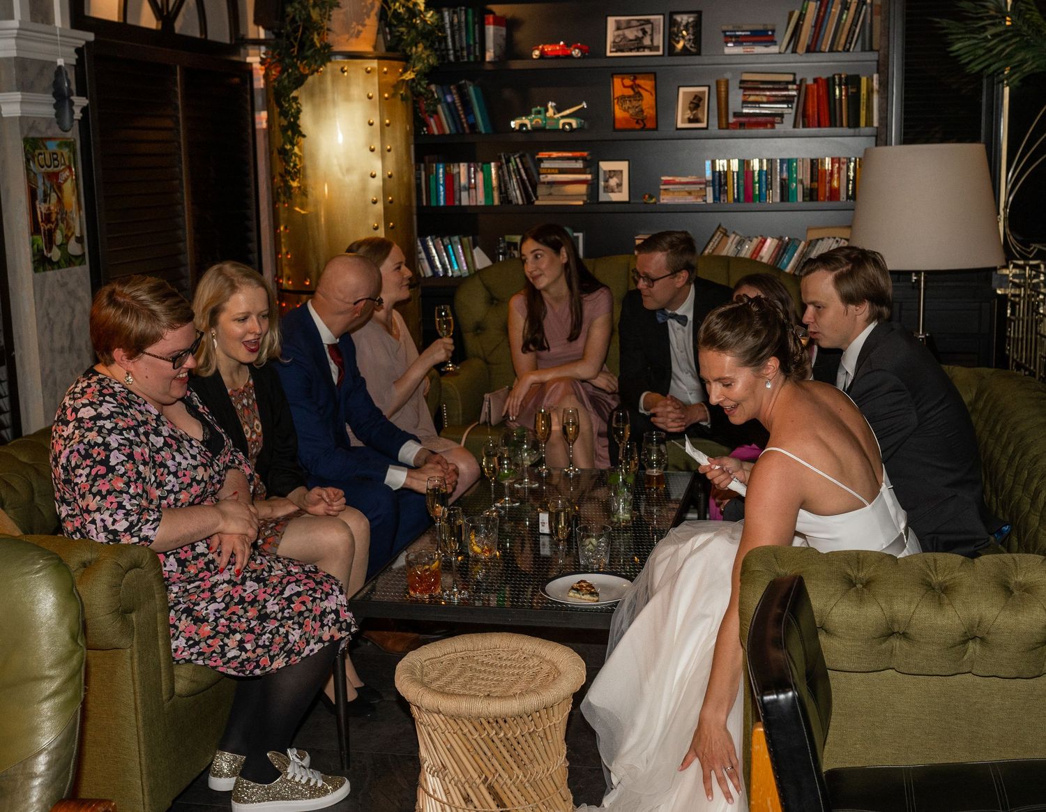 Wedding guests and bride in white dress socializing on green sofas in stylish venue.