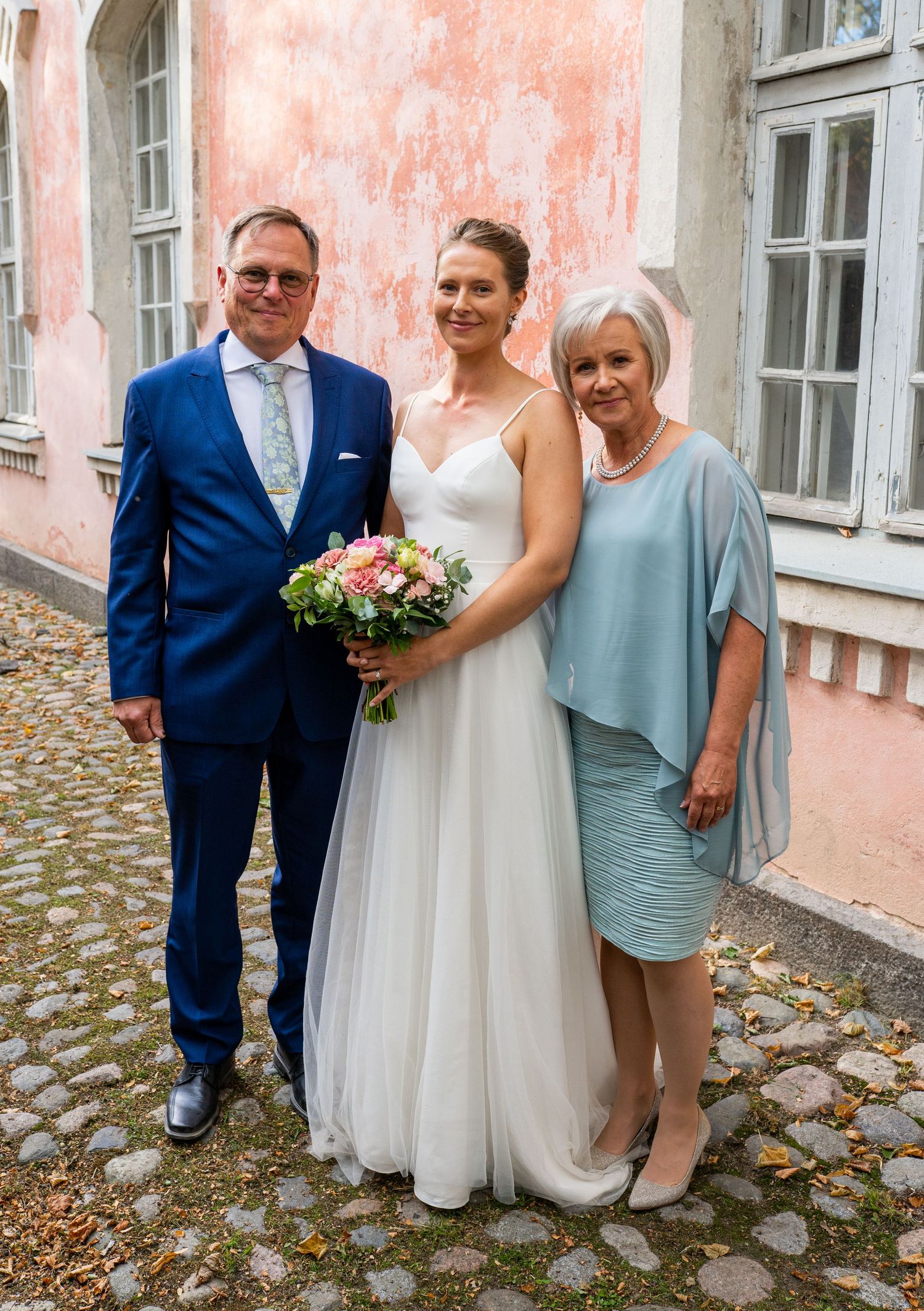 Bride in white tulle gown poses with bouquet between parents in blue suit and light blue dress outdoors.