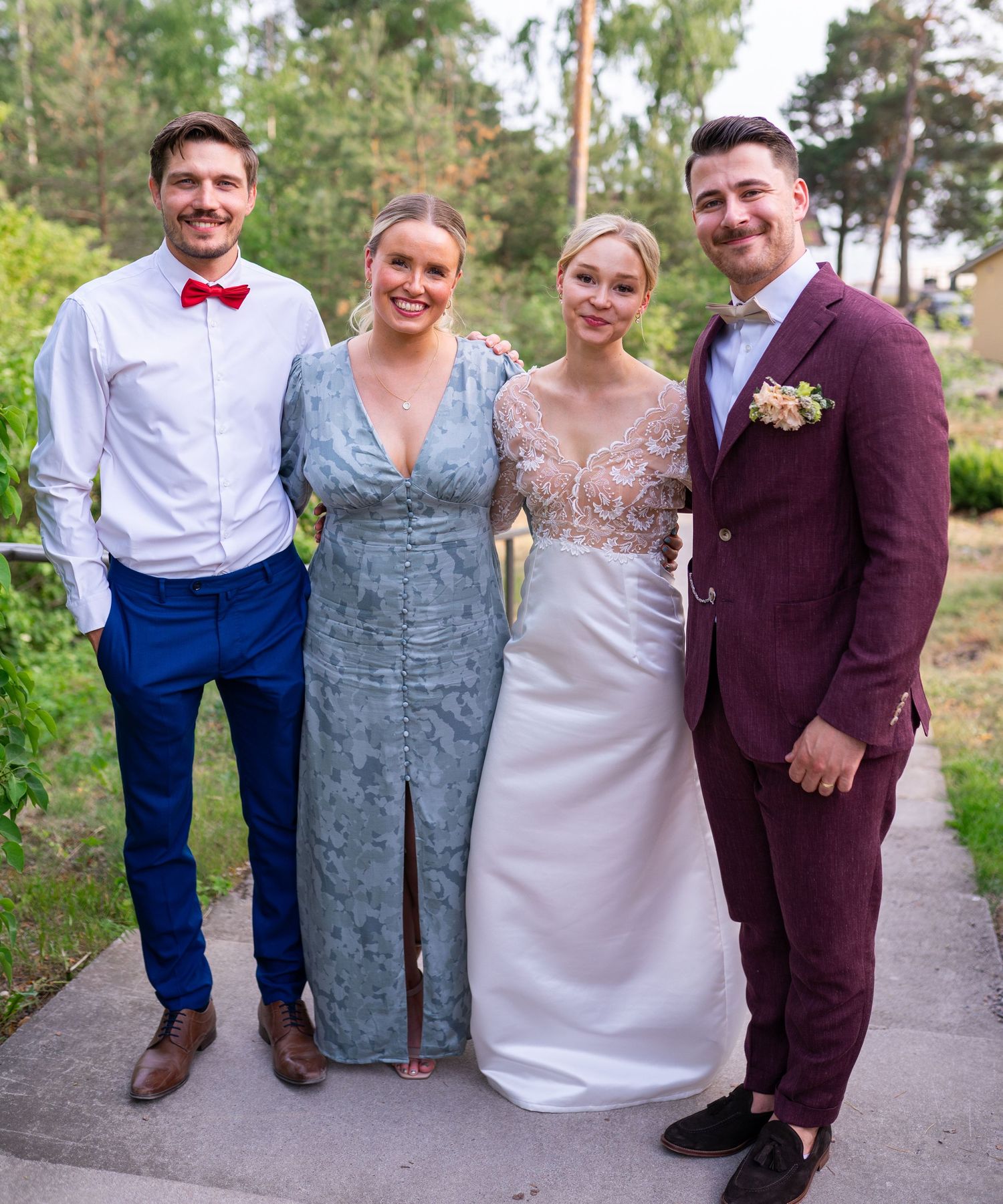 Four wedding guests pose together outdoors, including a bride in white lace and a groom in burgundy suit.
