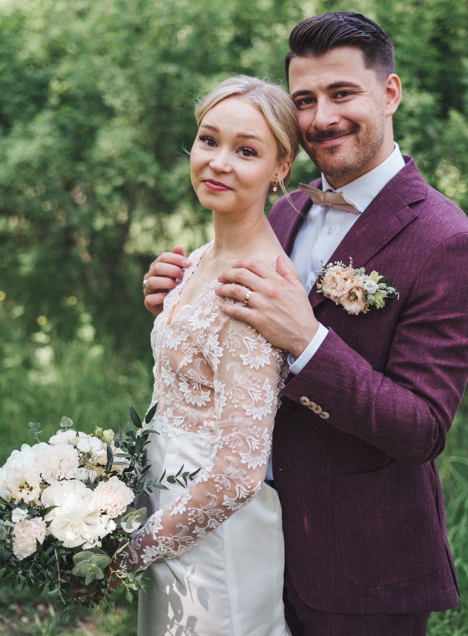 Bride and groom posing together outdoors, groom in burgundy suit embracing bride in lace dress with bouquet.