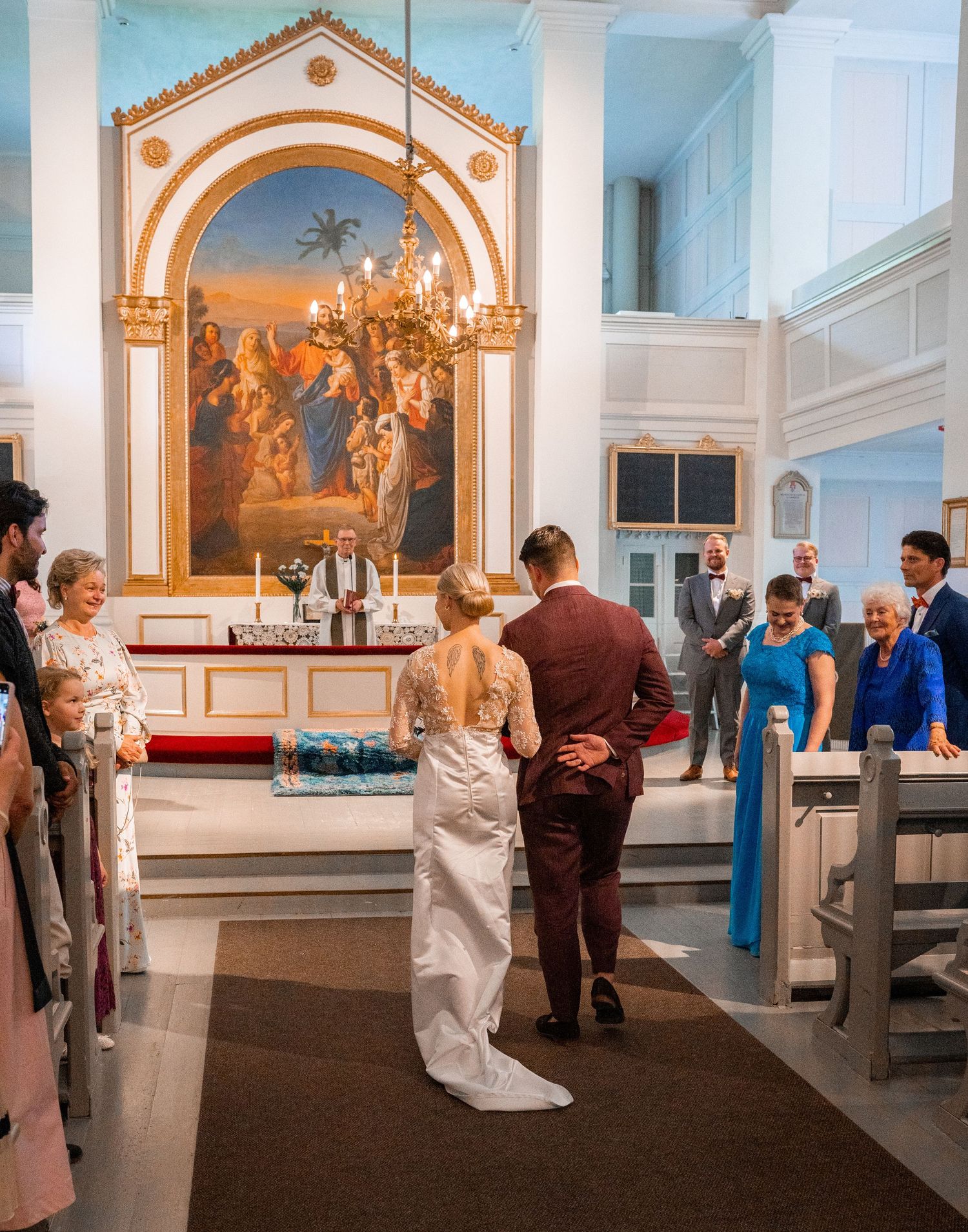 Bride and groom walk toward the altar in a white church with a ornate golden altarpiece painting during wedding.