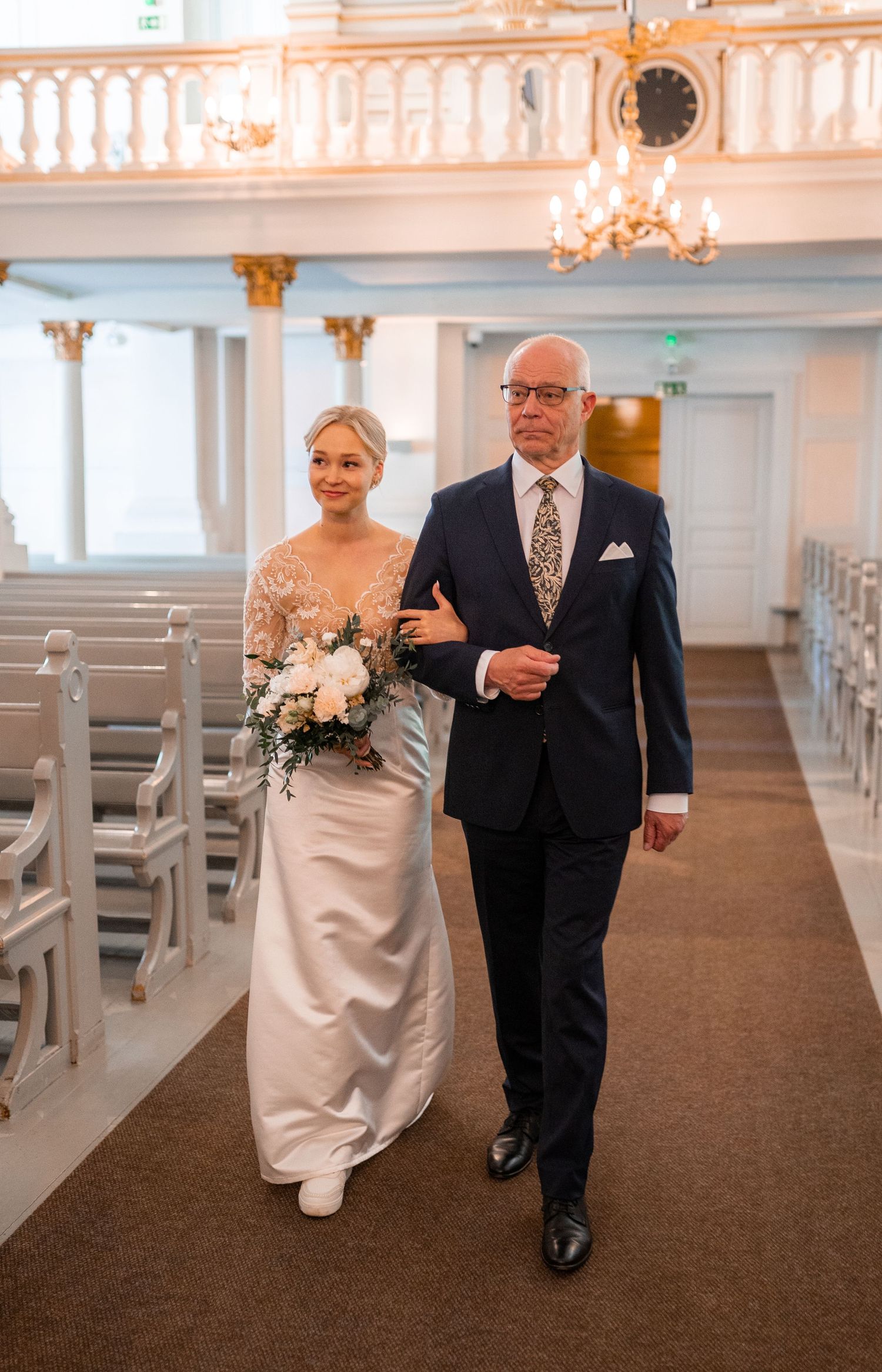 Bride in white lace gown holding floral bouquet walks down church aisle with escort in navy suit.