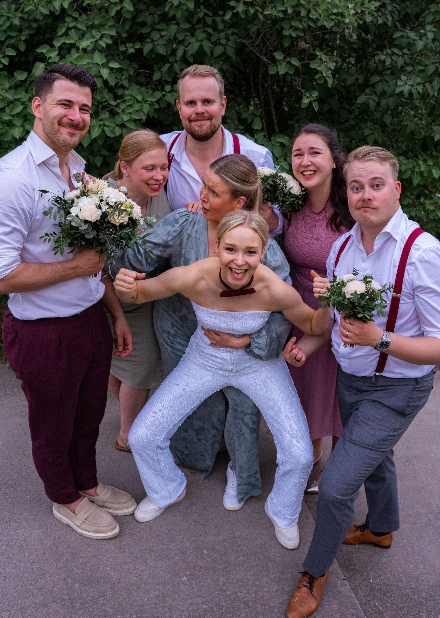 Bride being lifted by wedding party in fun group photo outdoors with white floral bouquets.