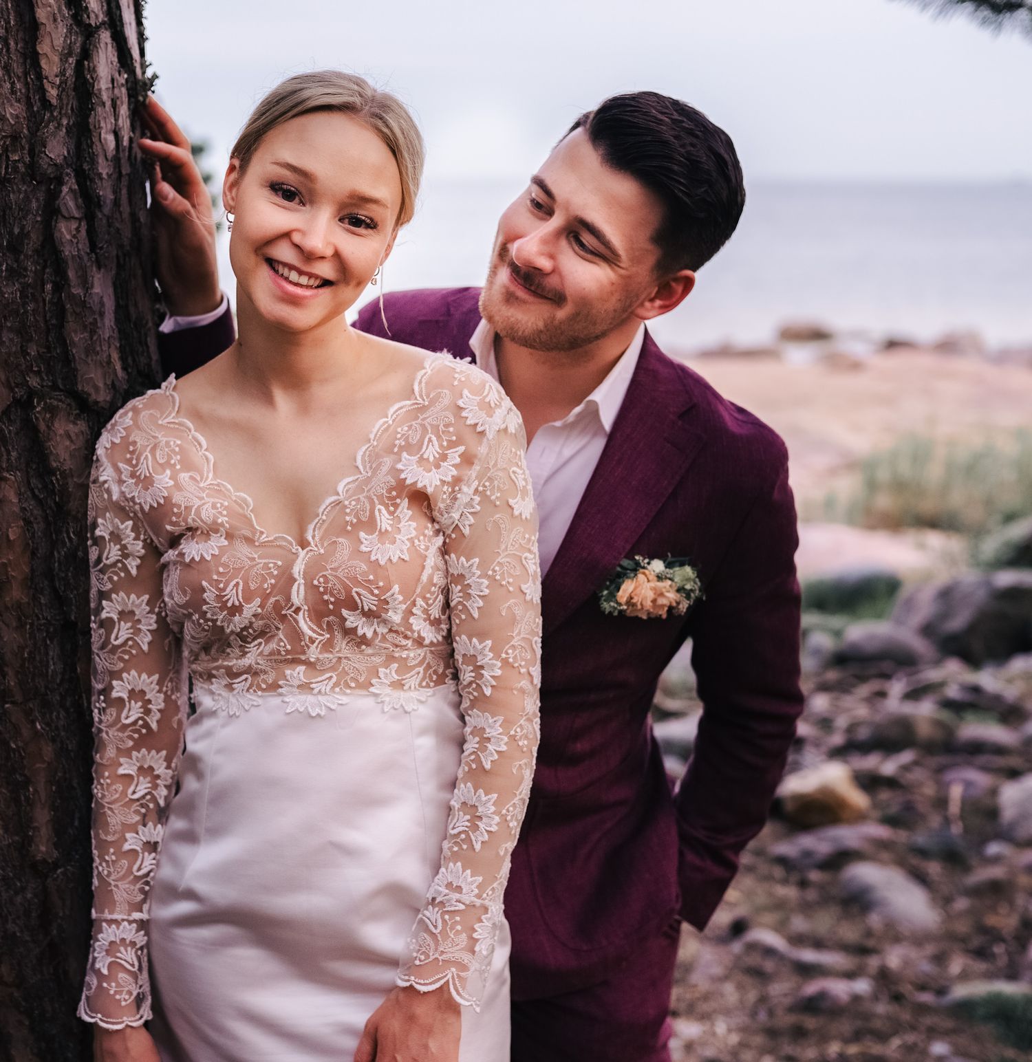 Smiling bride in lace wedding gown and groom in burgundy suit posing together by a tree near the sea.