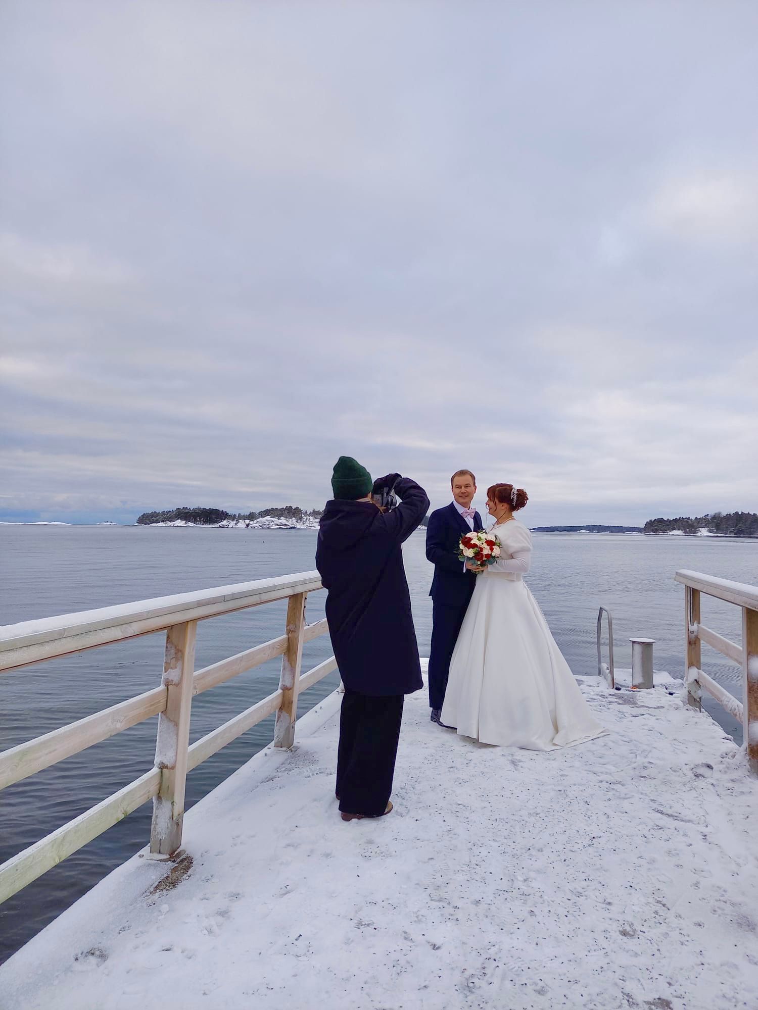 Wedding photographer shoots couple on snowy waterfront pier under cloudy winter sky.