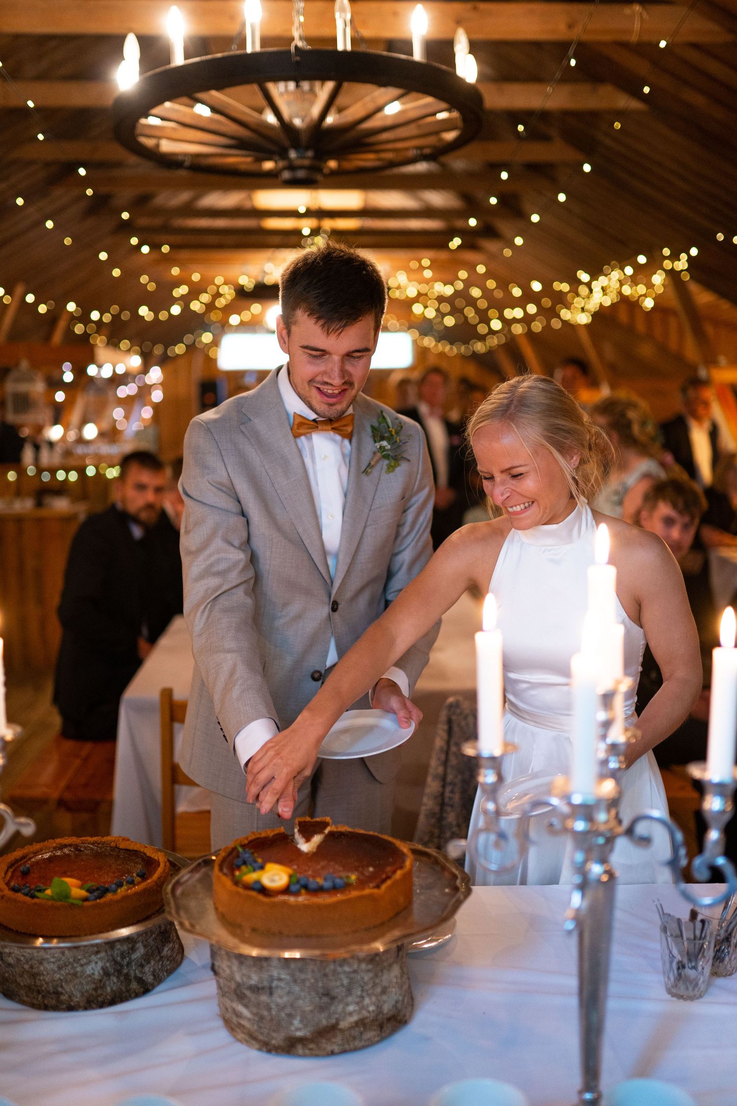 Smiling newlyweds cut their wedding cake in a warmly lit barn reception venue.