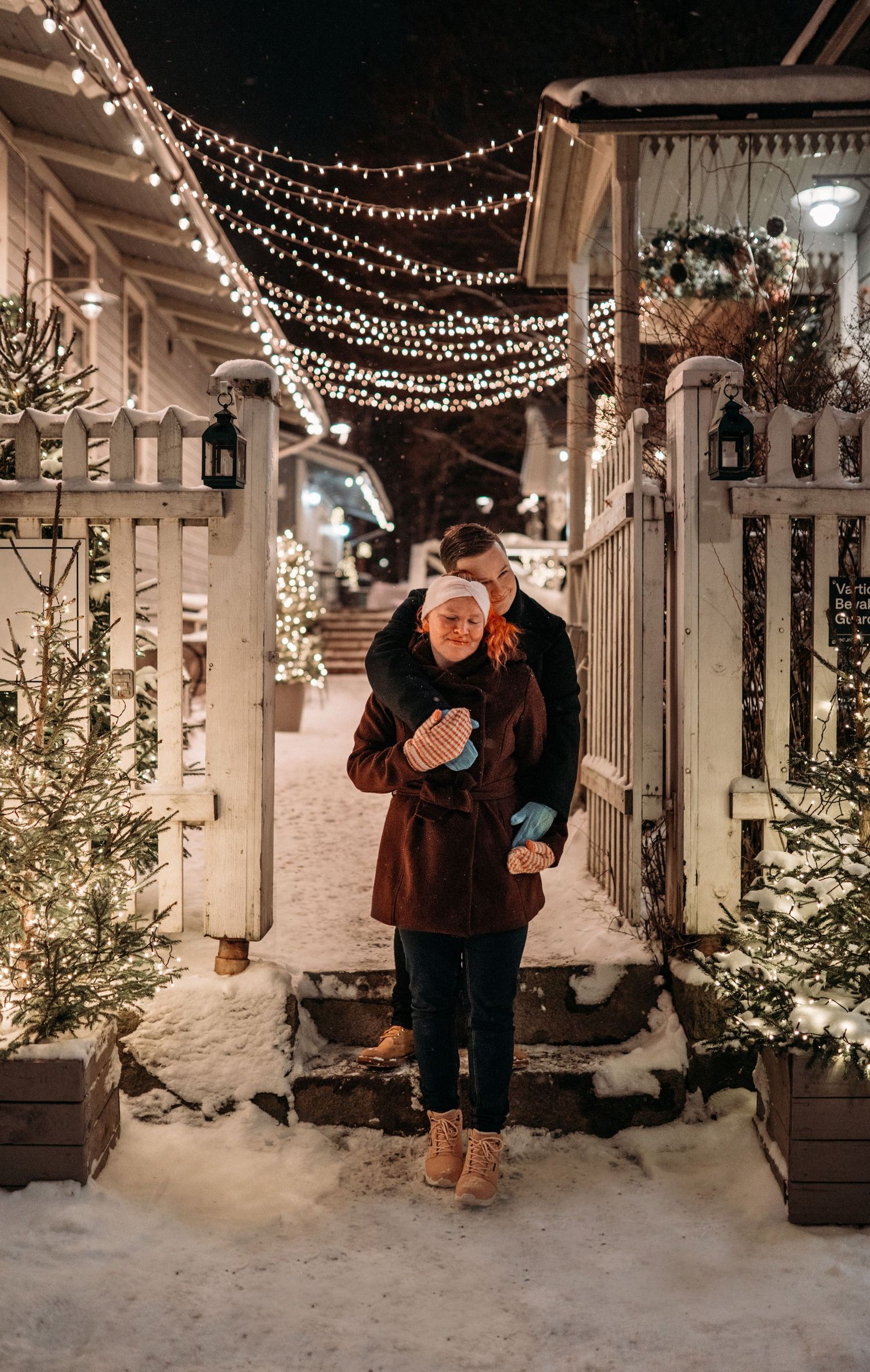 Couple embracing under festive Christmas lights in snowy outdoor market at night.