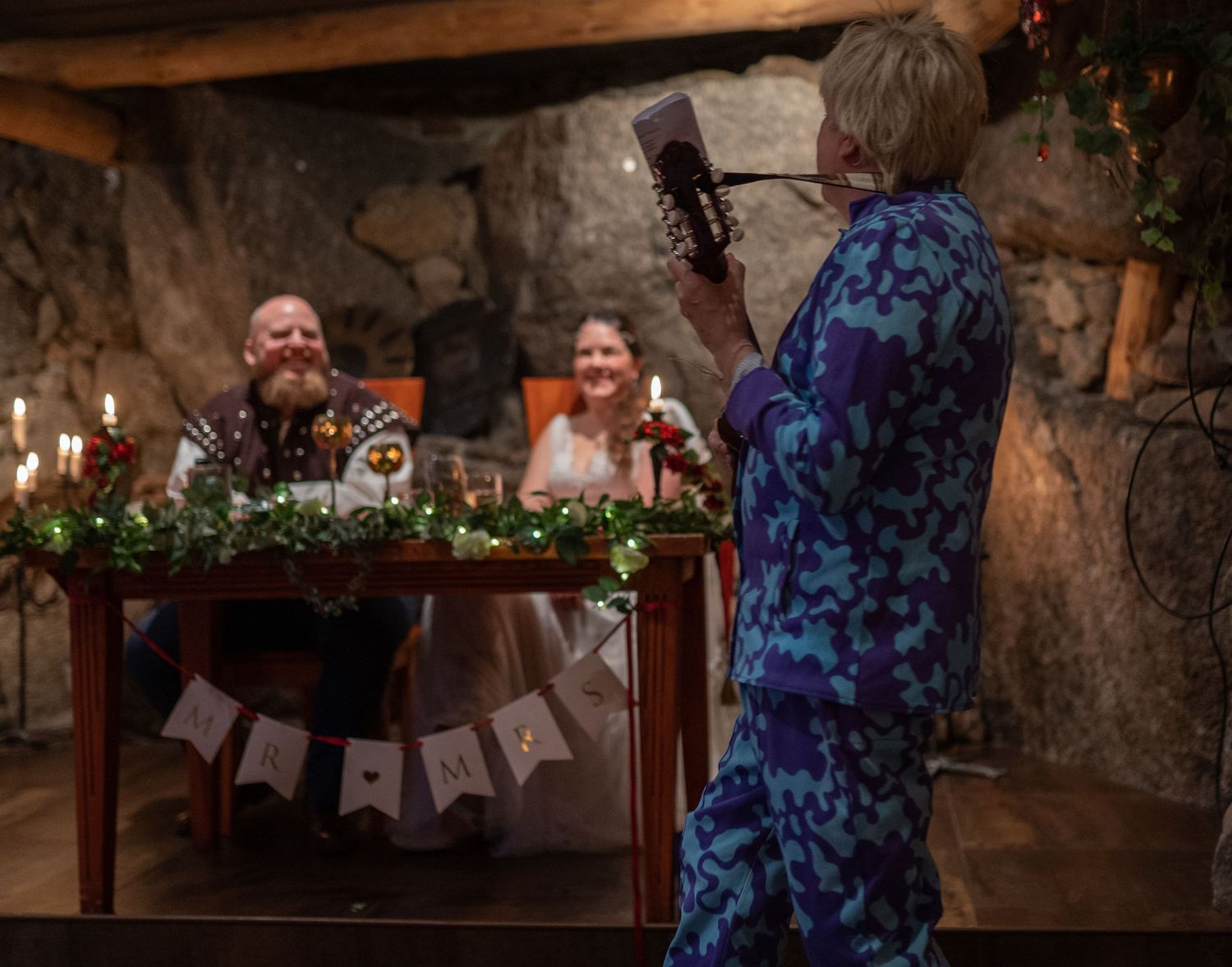 Wedding couple seated at decorated table while officiant reads from book in rustic stone venue with candles.