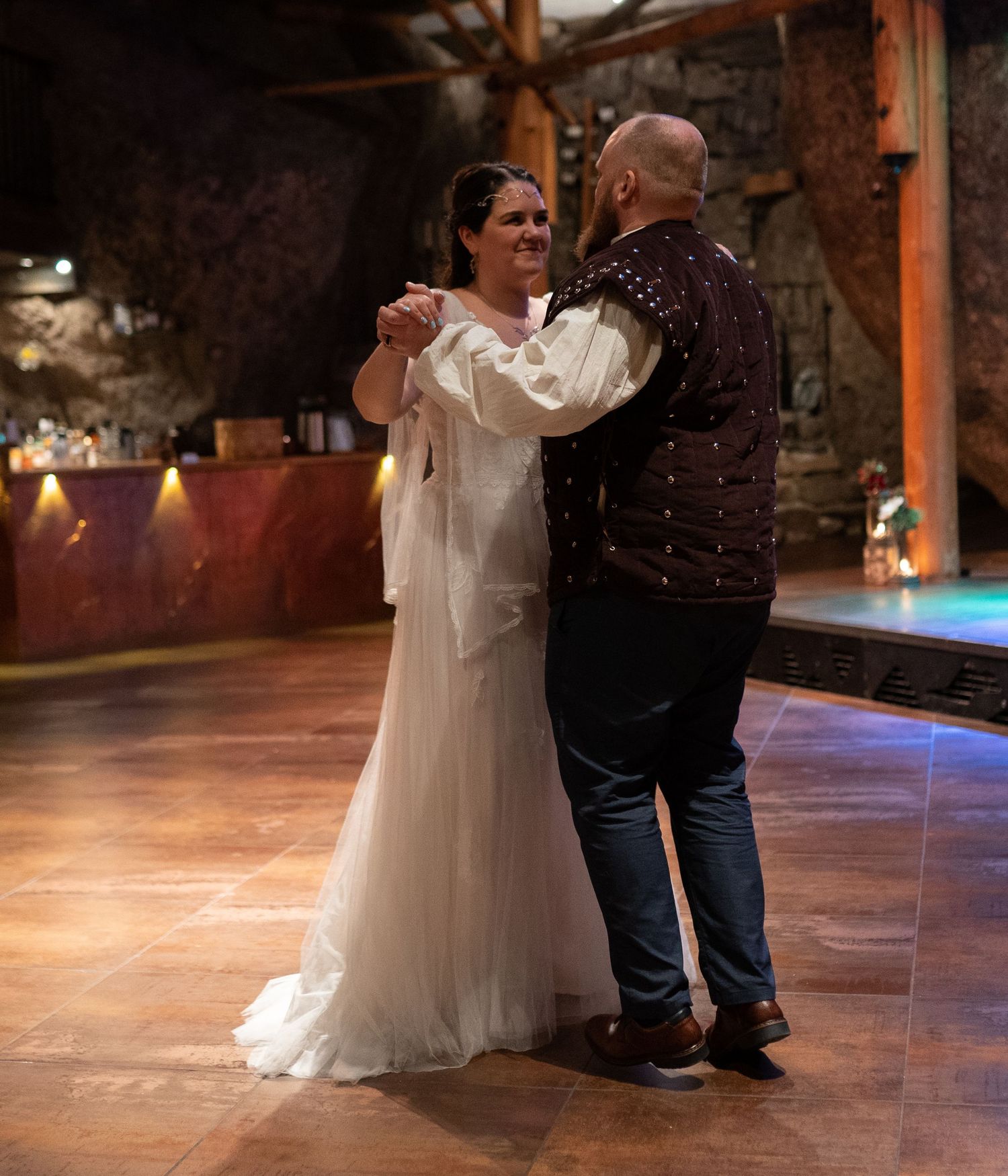 Couple sharing first dance in wedding attire inside a rustic stone venue with warm ambient lighting.
