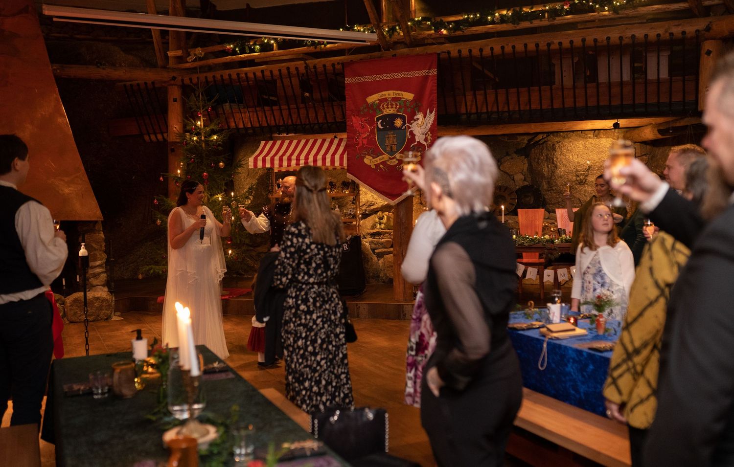 Medieval banquet hall gathering with guests in period costumes, candles, and a red heraldic banner displayed above.