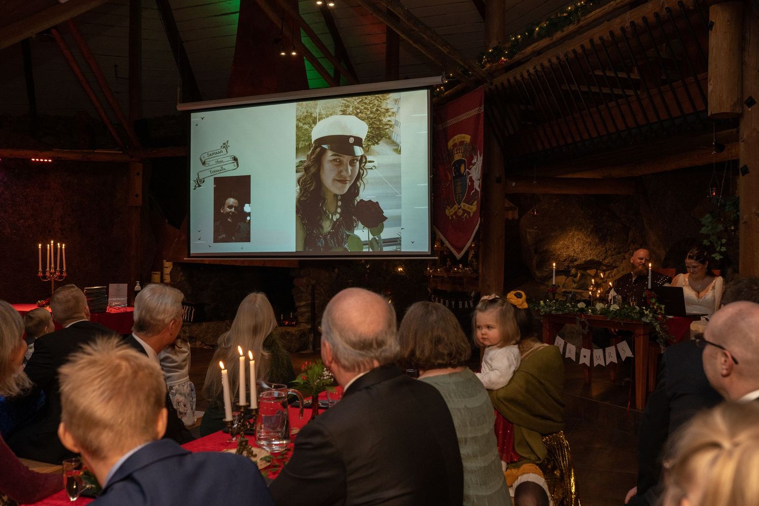Guests at a festive dinner event watch a projected photo of a woman on a large screen in a decorated hall.