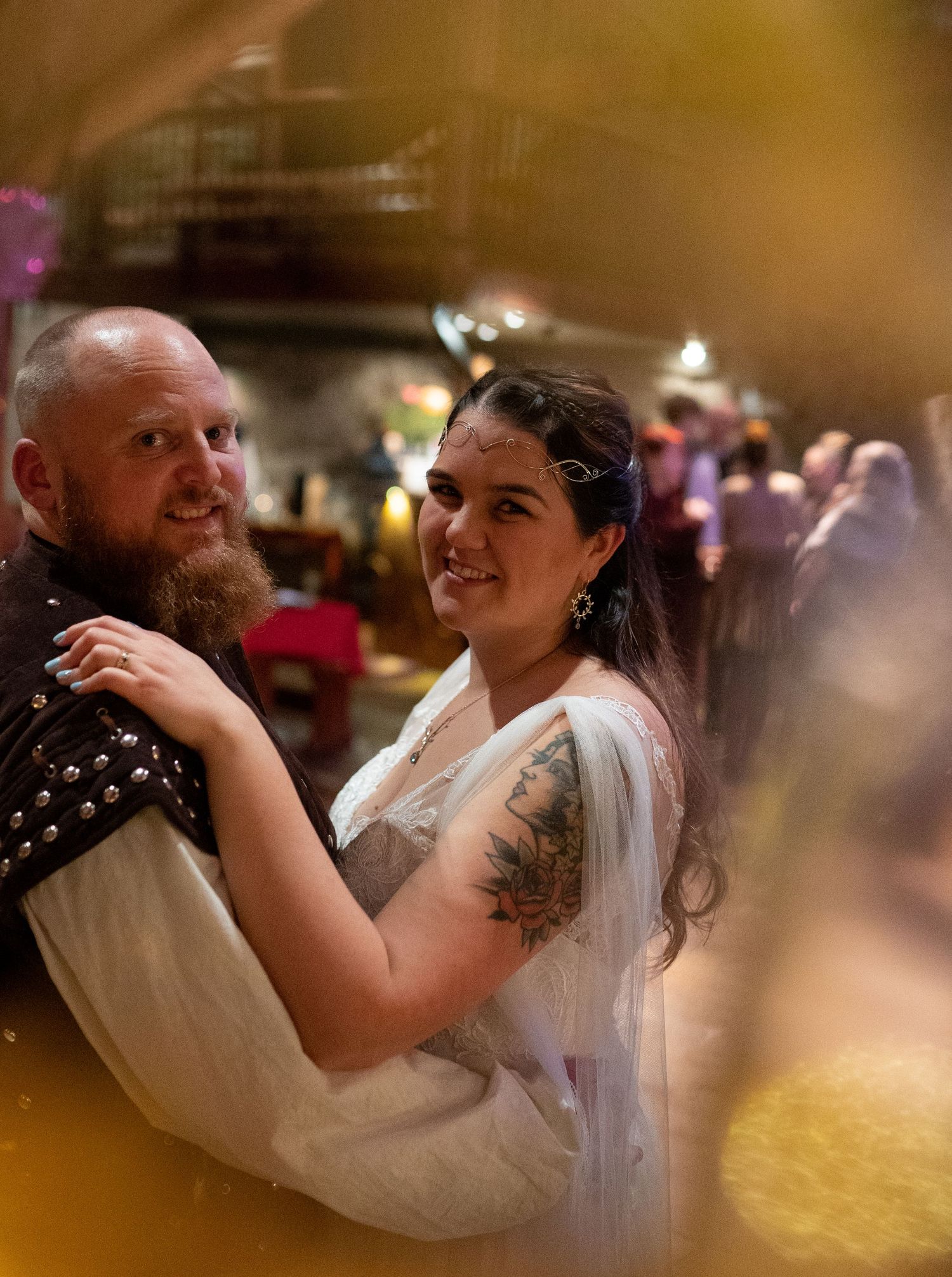 A bearded groom in medieval attire smiles with his tattooed bride in a white dress and tiara at a wedding reception.