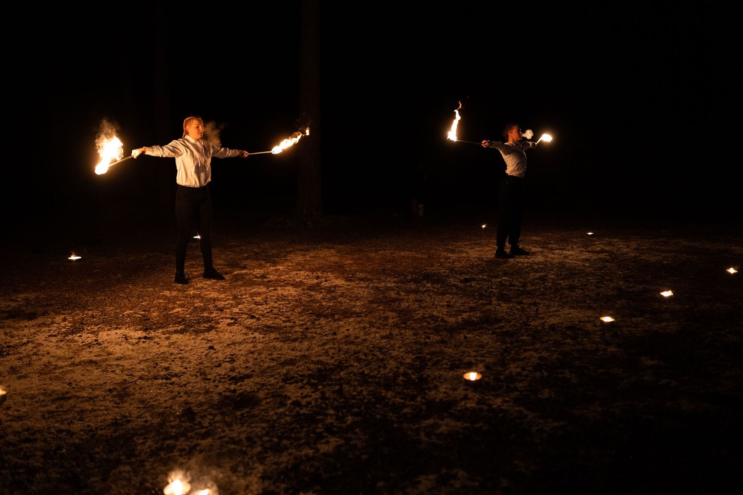Two fire dancers perform at night, holding flaming torches with arms outstretched, surrounded by small ground lights.