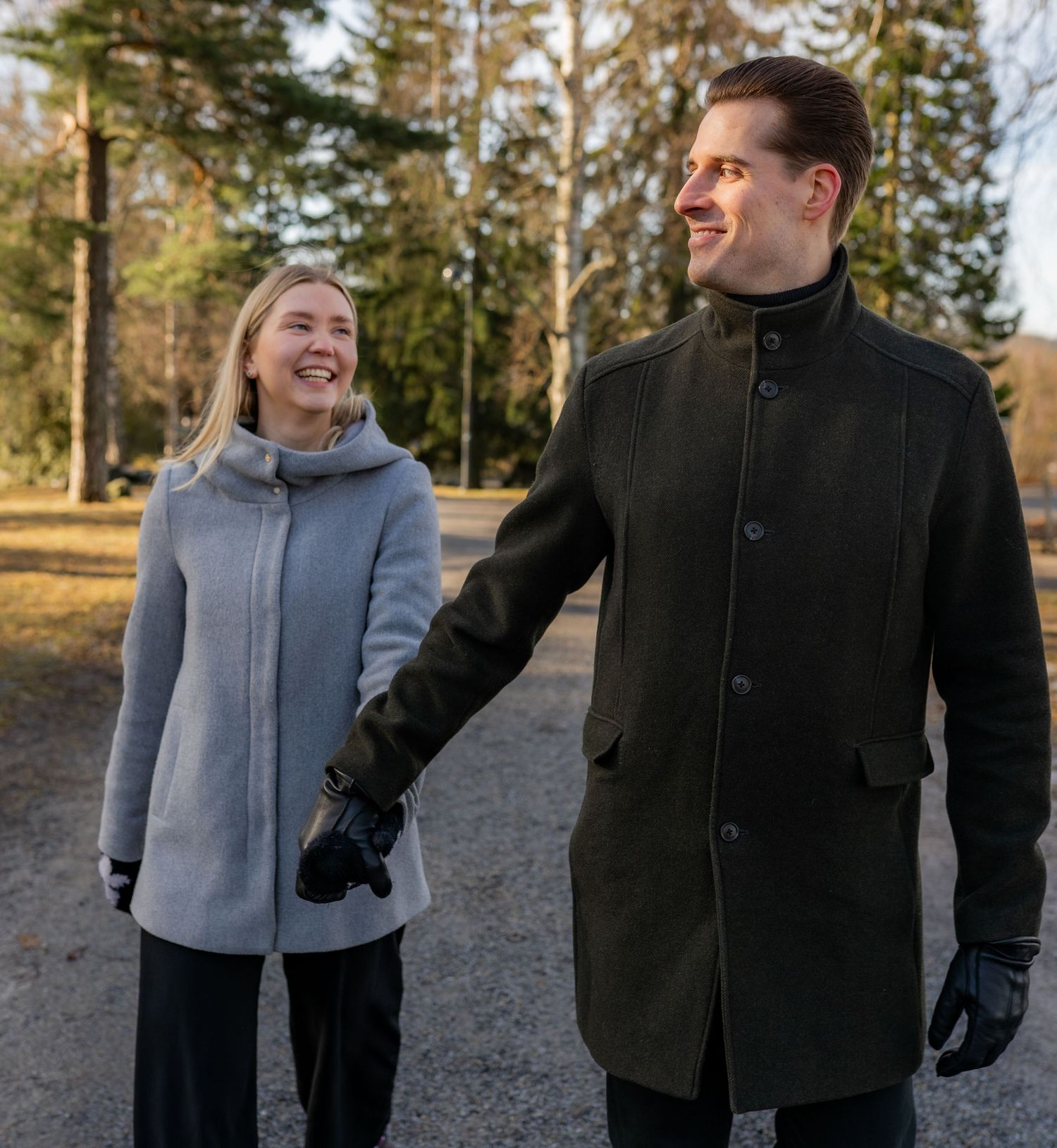 Couple in winter coats holding hands while walking outdoors on a gravel path surrounded by trees.