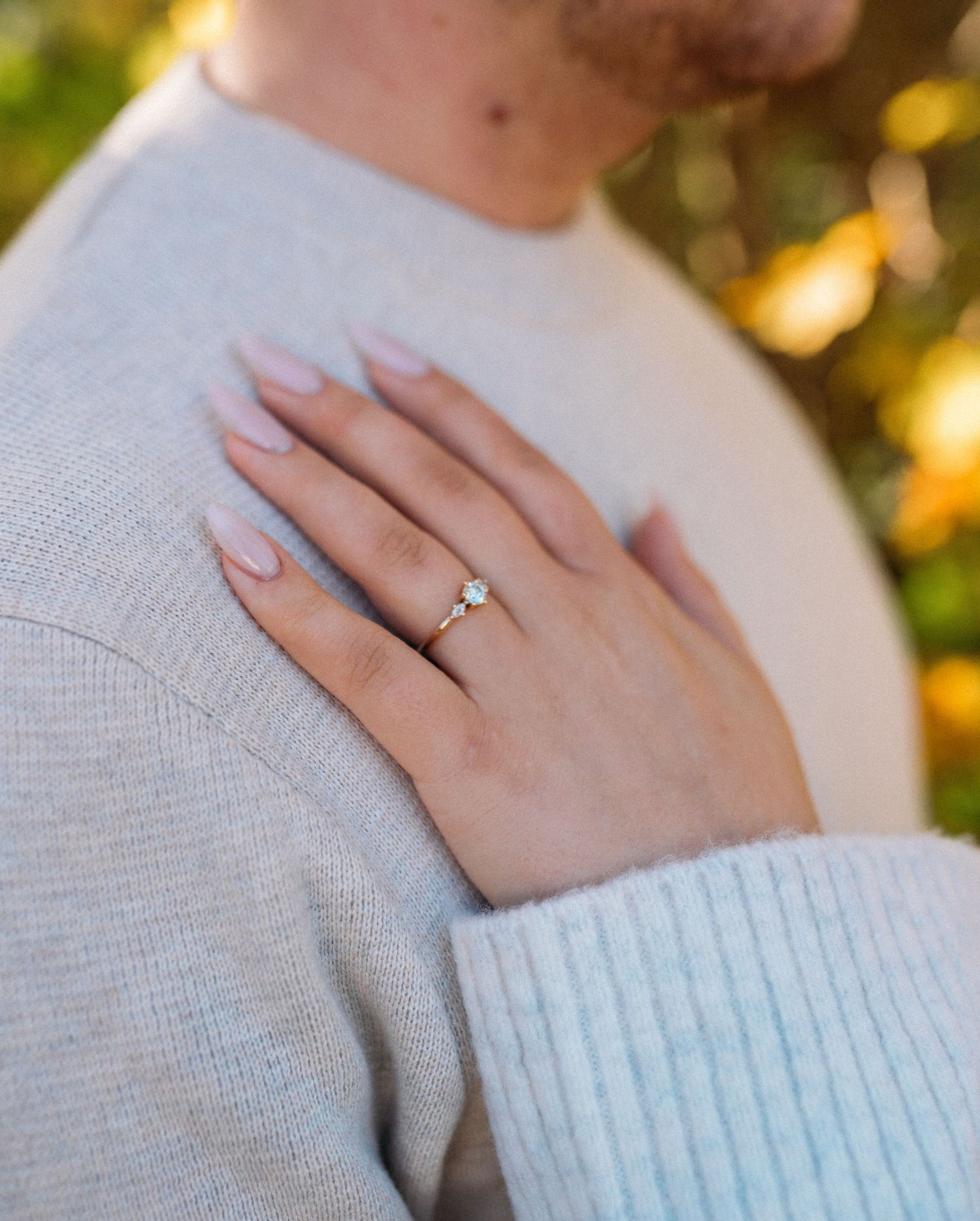 Close-up of a woman's hand with a diamond solitaire engagement ring on a gold band, resting on a man's shoulder.