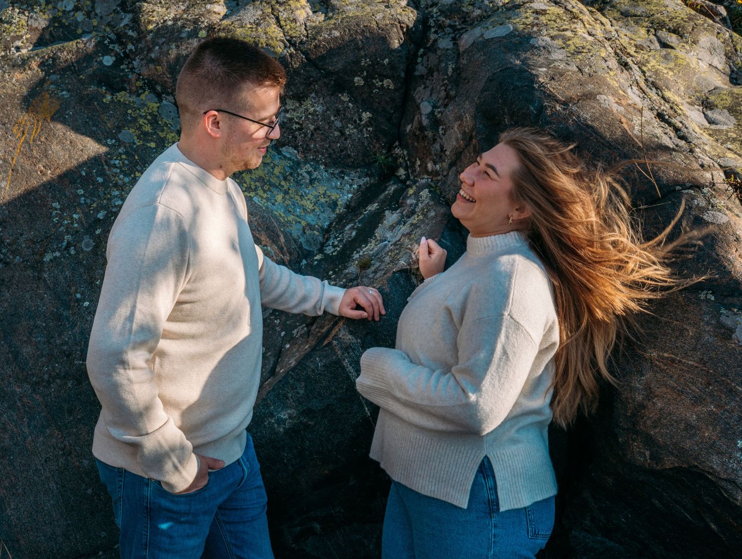 Couple holding hands near rocky cliff, woman's hair blowing in wind, both wearing cream sweaters and jeans.