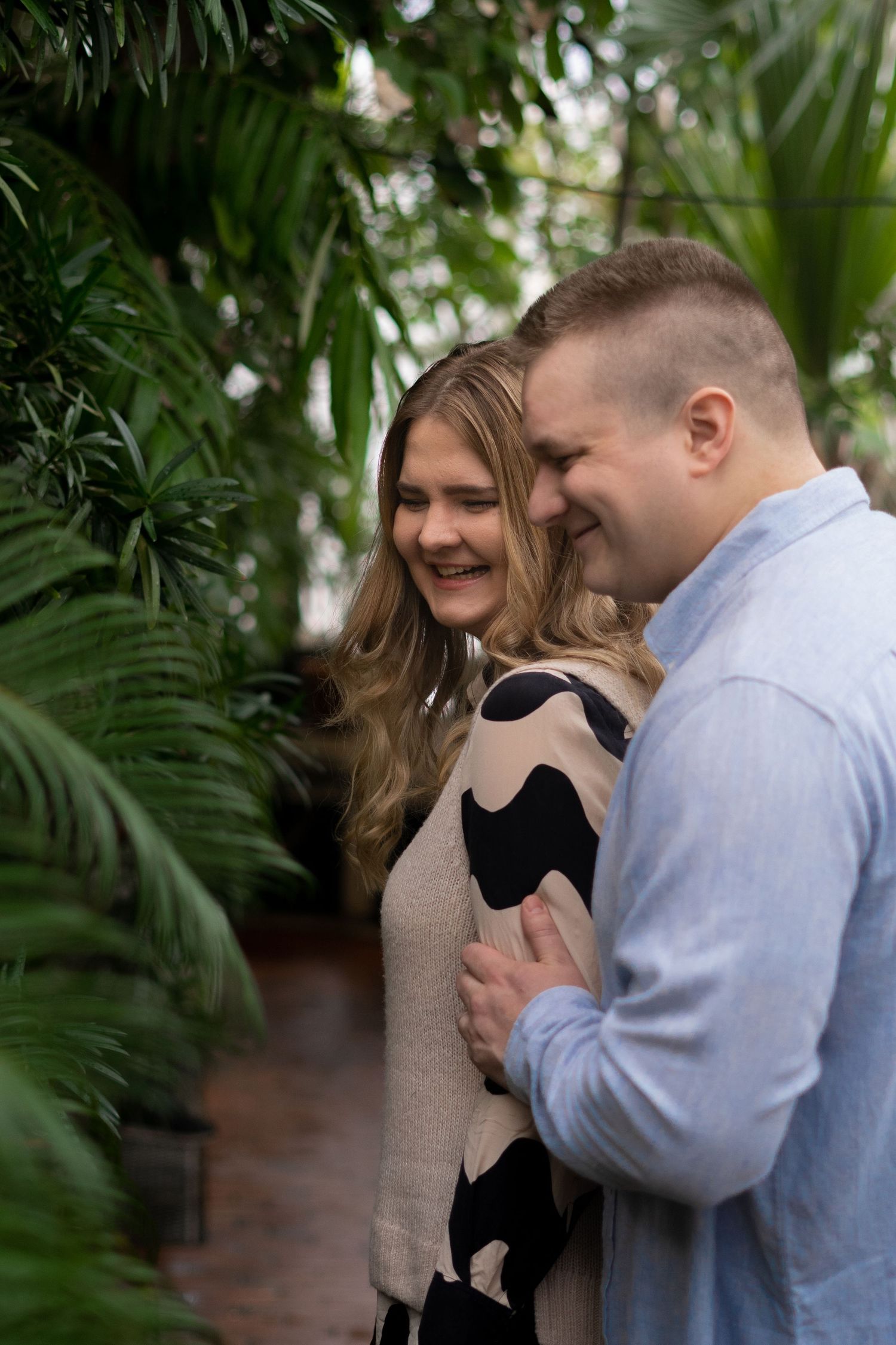 A smiling couple embraces among lush tropical plants, the woman in a patterned dress and man in a blue shirt.