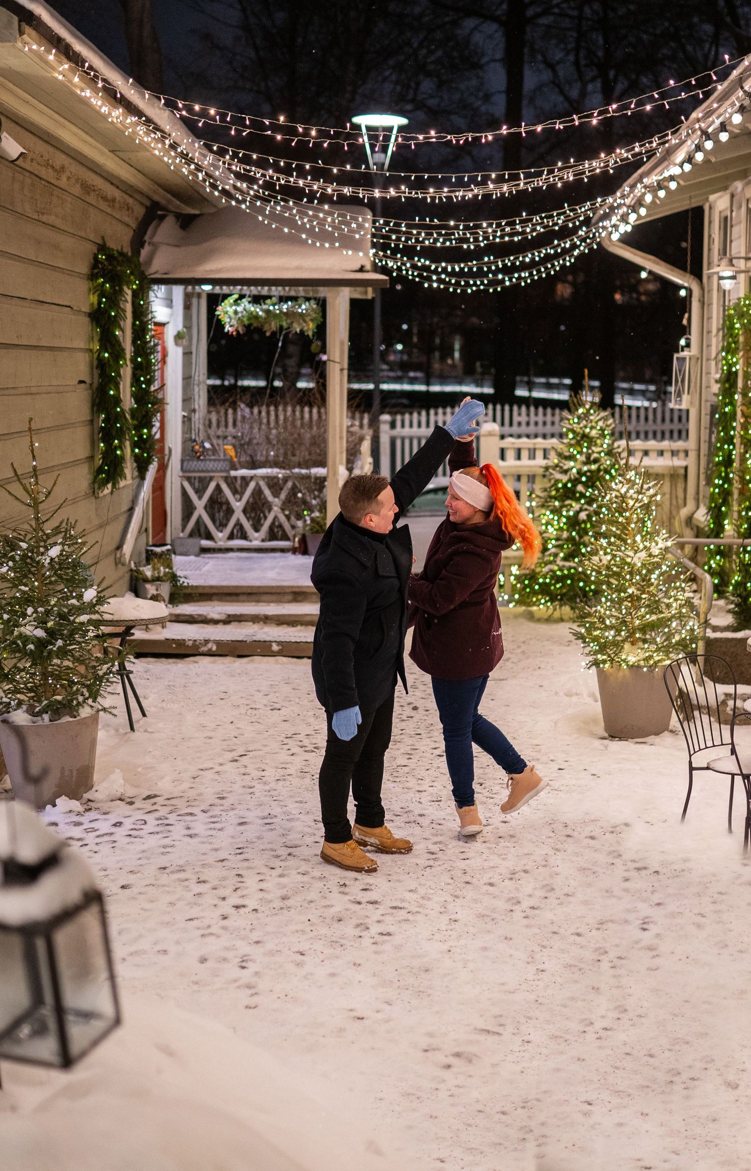 Couple dancing outdoors in snow at night surrounded by festive Christmas lights and decorated trees.