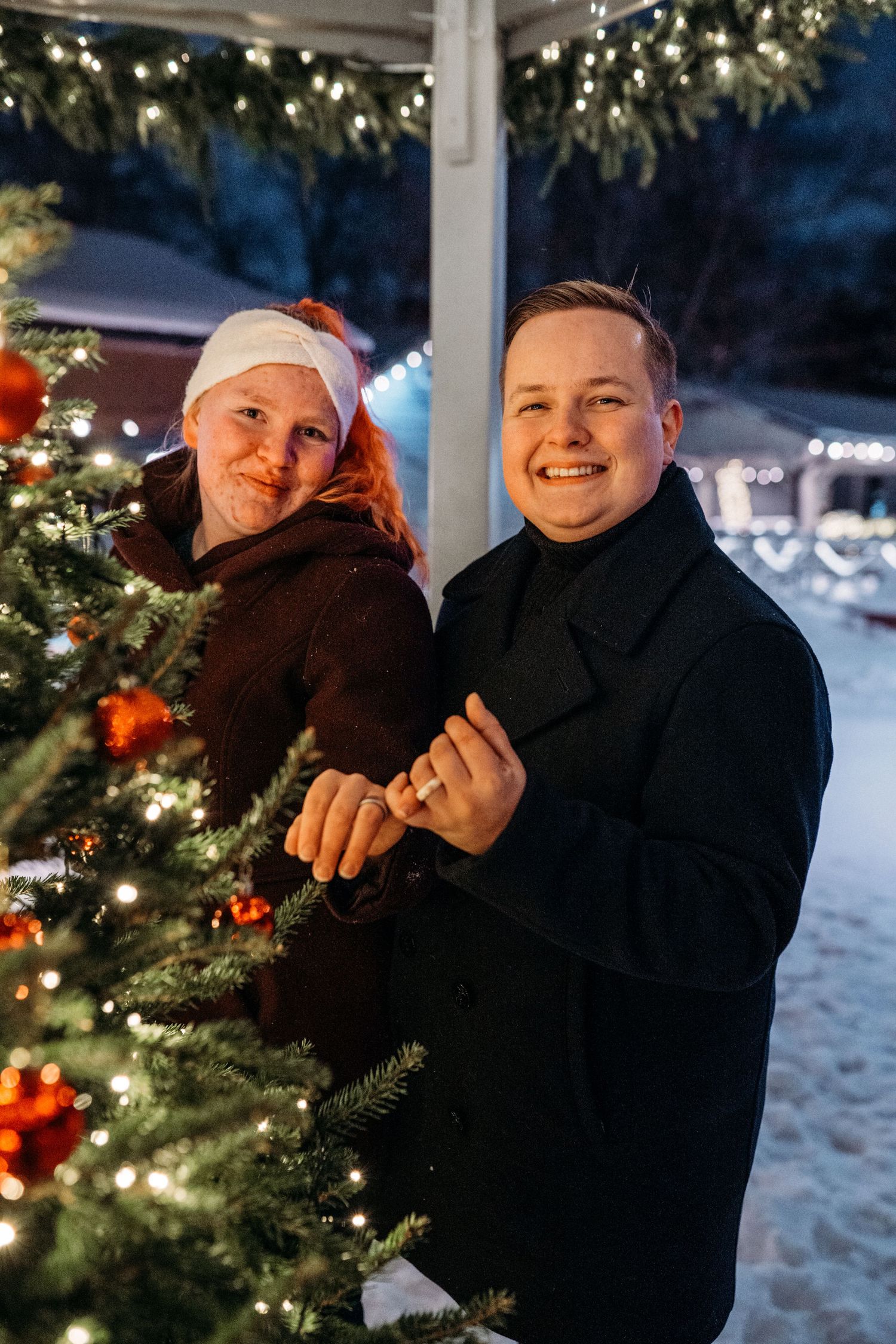 Couple smiling by a decorated Christmas tree with red ornaments and lights outdoors at night in winter.