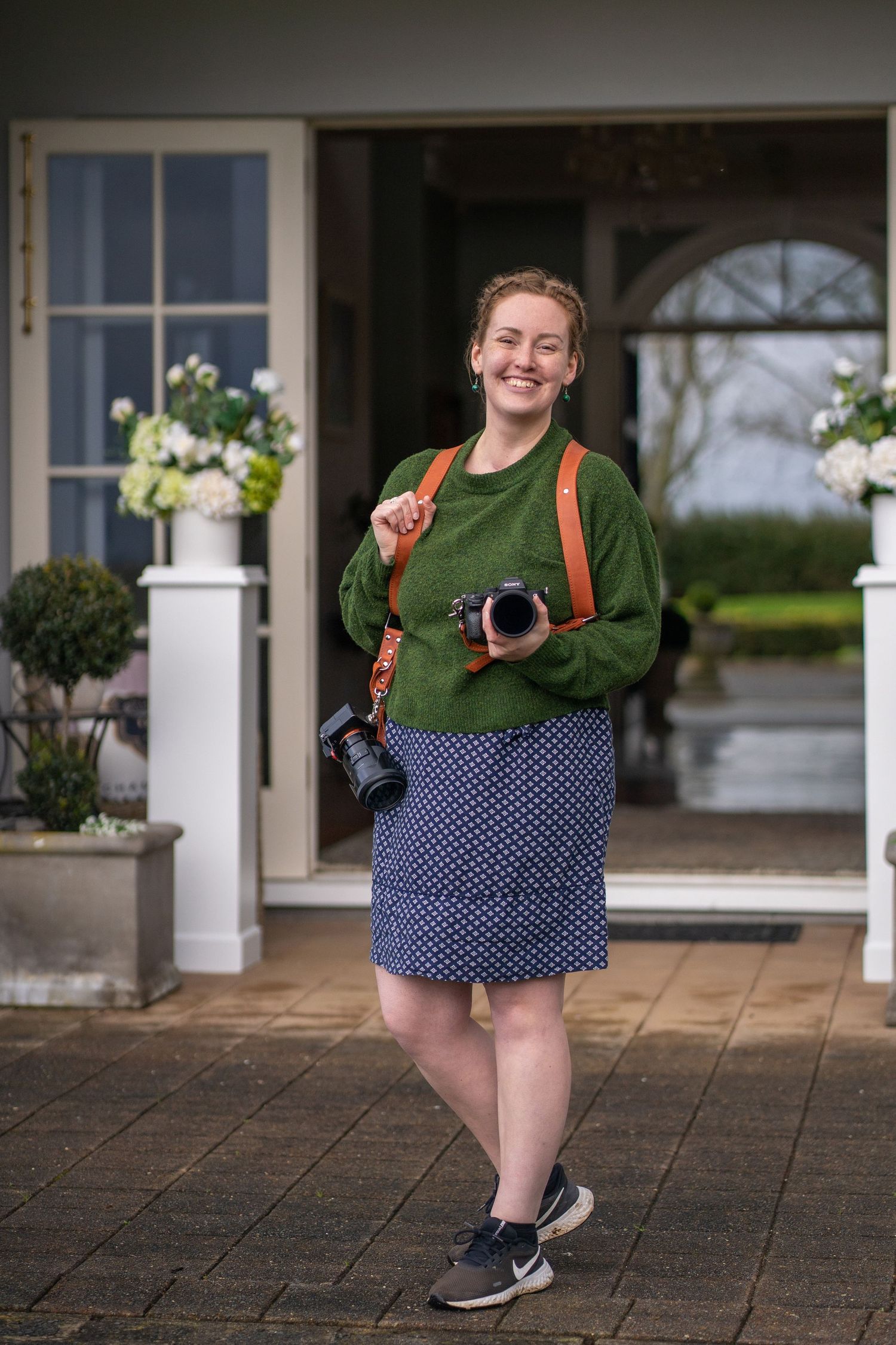 Smiling female photographer holding cameras, wearing green sweater and polka dot skirt, standing outside elegant white building.