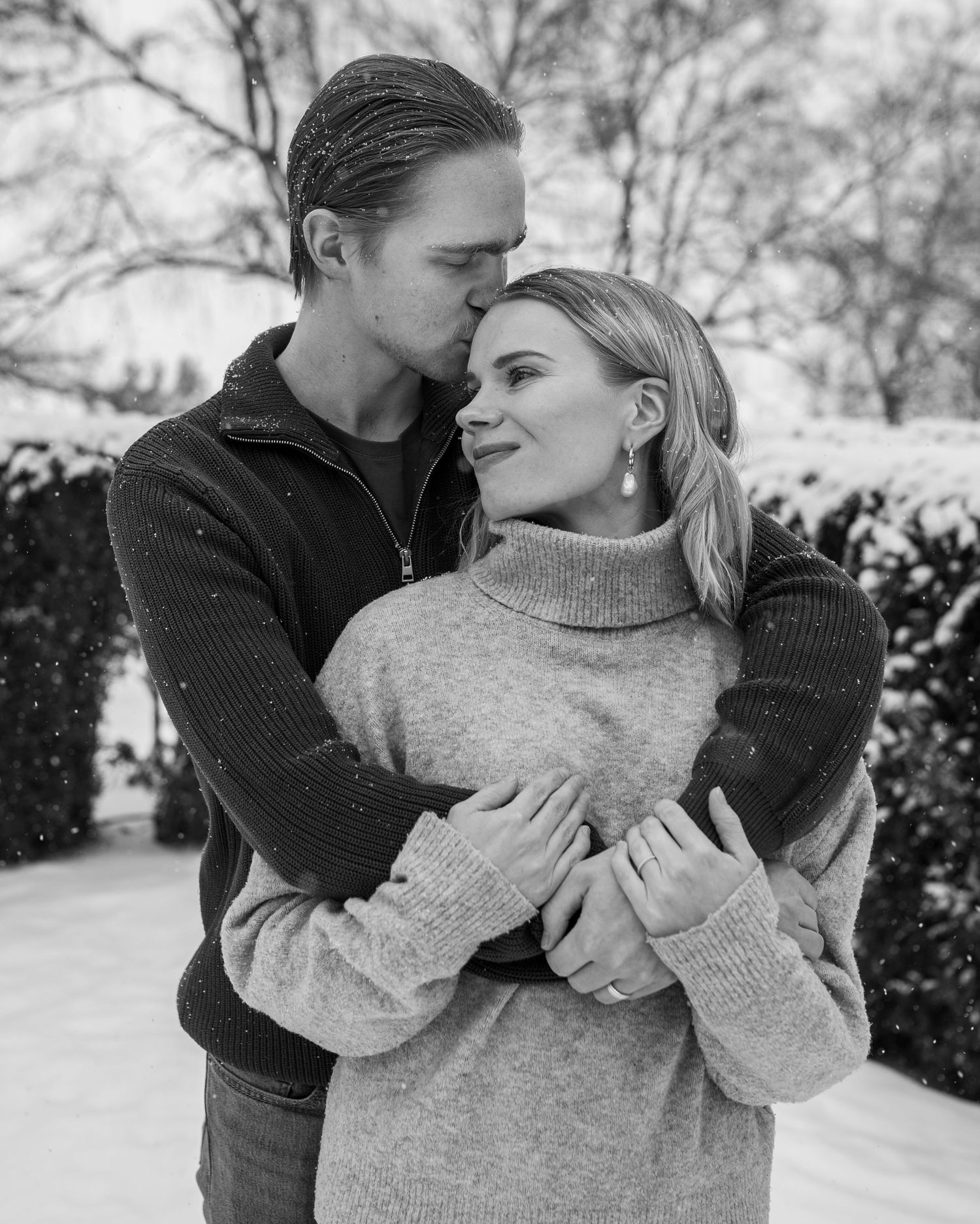 Black and white photo of a couple embracing in snowy winter setting, man kissing womans forehead tenderly.