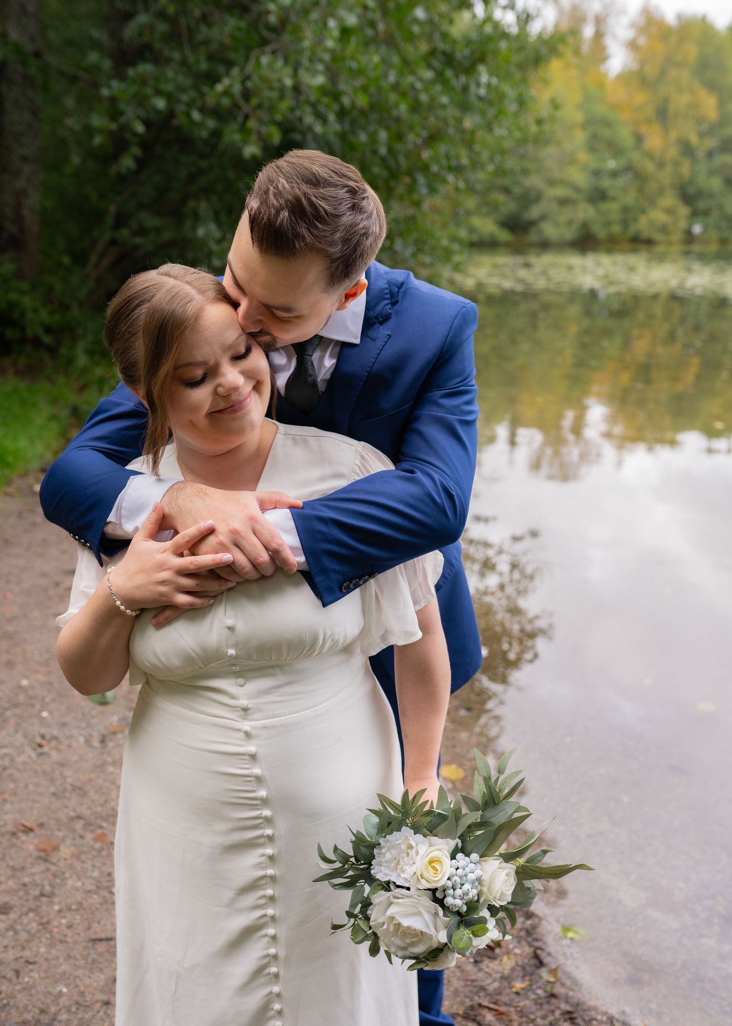 Groom in blue suit embracing bride in white dress by a tranquil lakeside holding a bouquet.