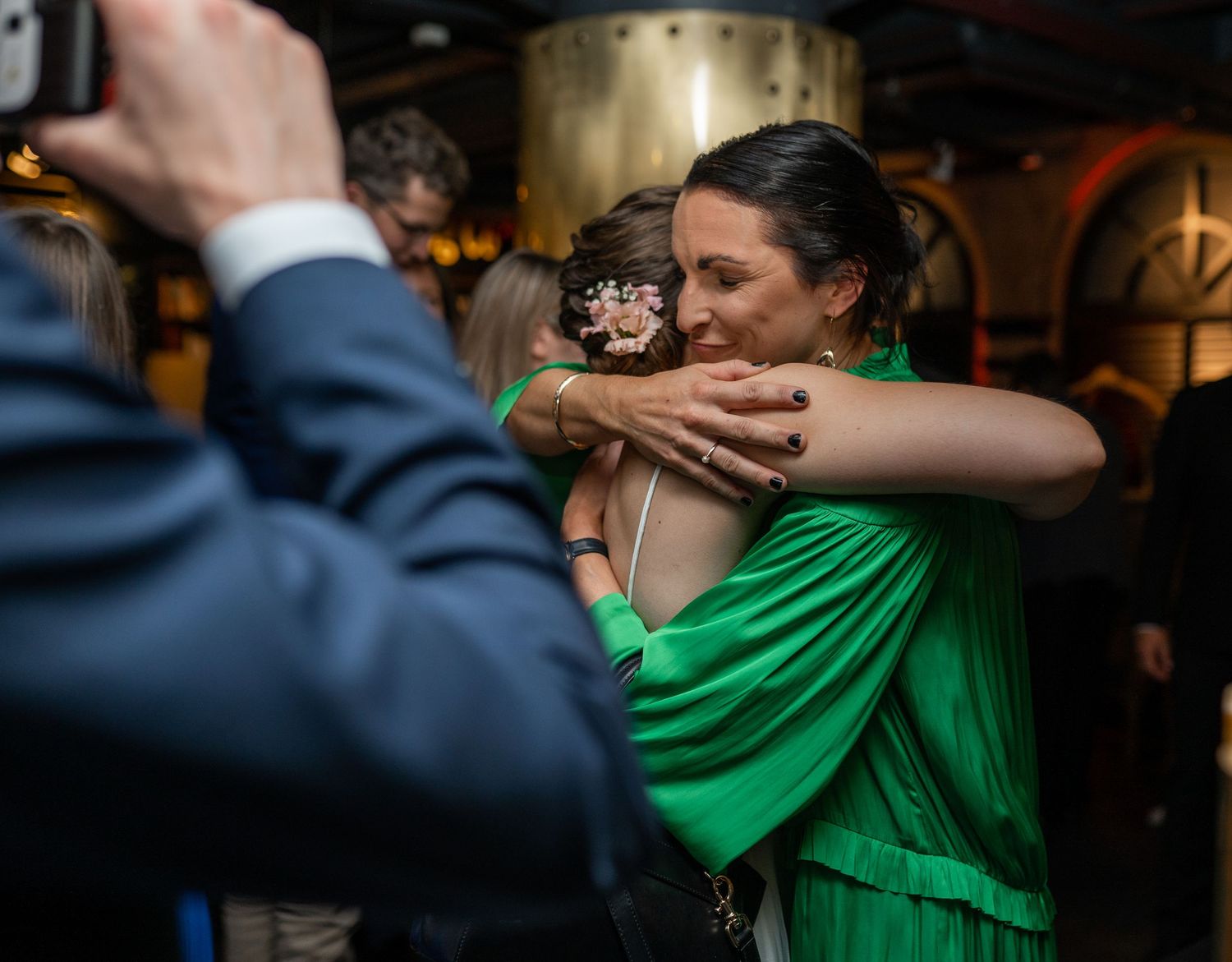 Bride in white dress receives warm hug from woman in green dress at wedding reception celebration.