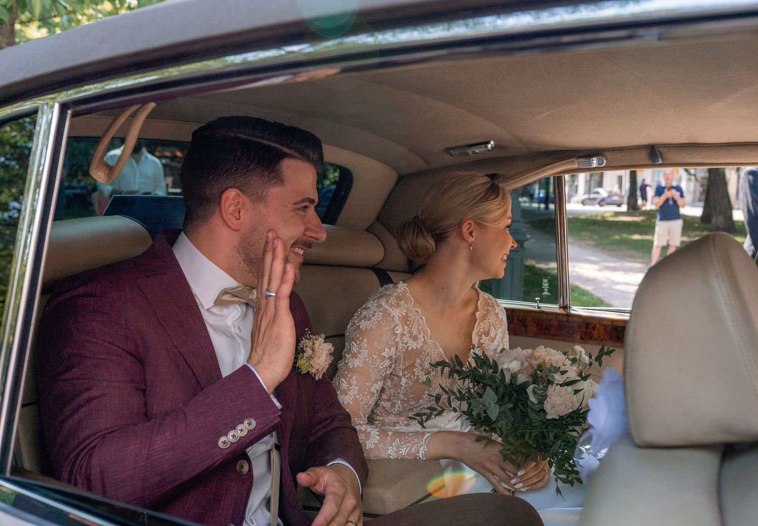 Groom gestures emotionally while sitting beside bouquet-holding bride inside elegant vintage wedding car.