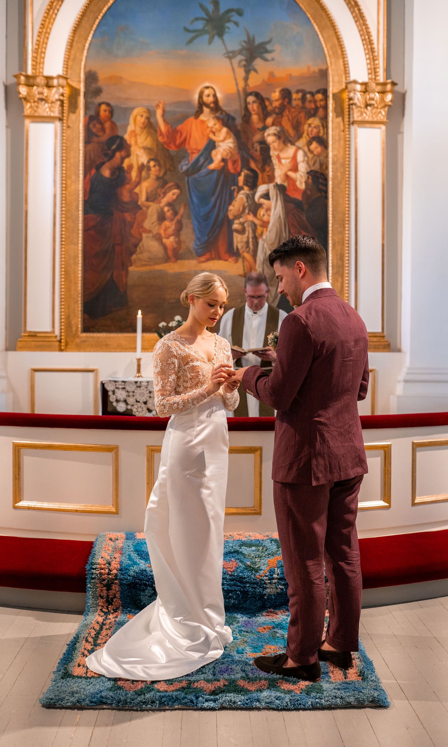 Bride in lace gown exchanges rings with groom before ornate golden altarpiece painting in church ceremony.