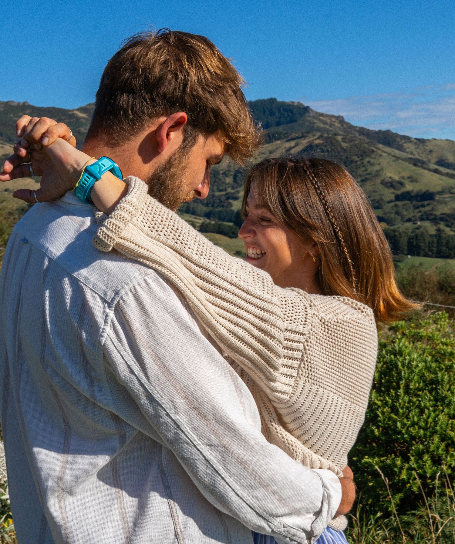 Couple shares joyful moment together on hillside with green mountains under clear blue sky.