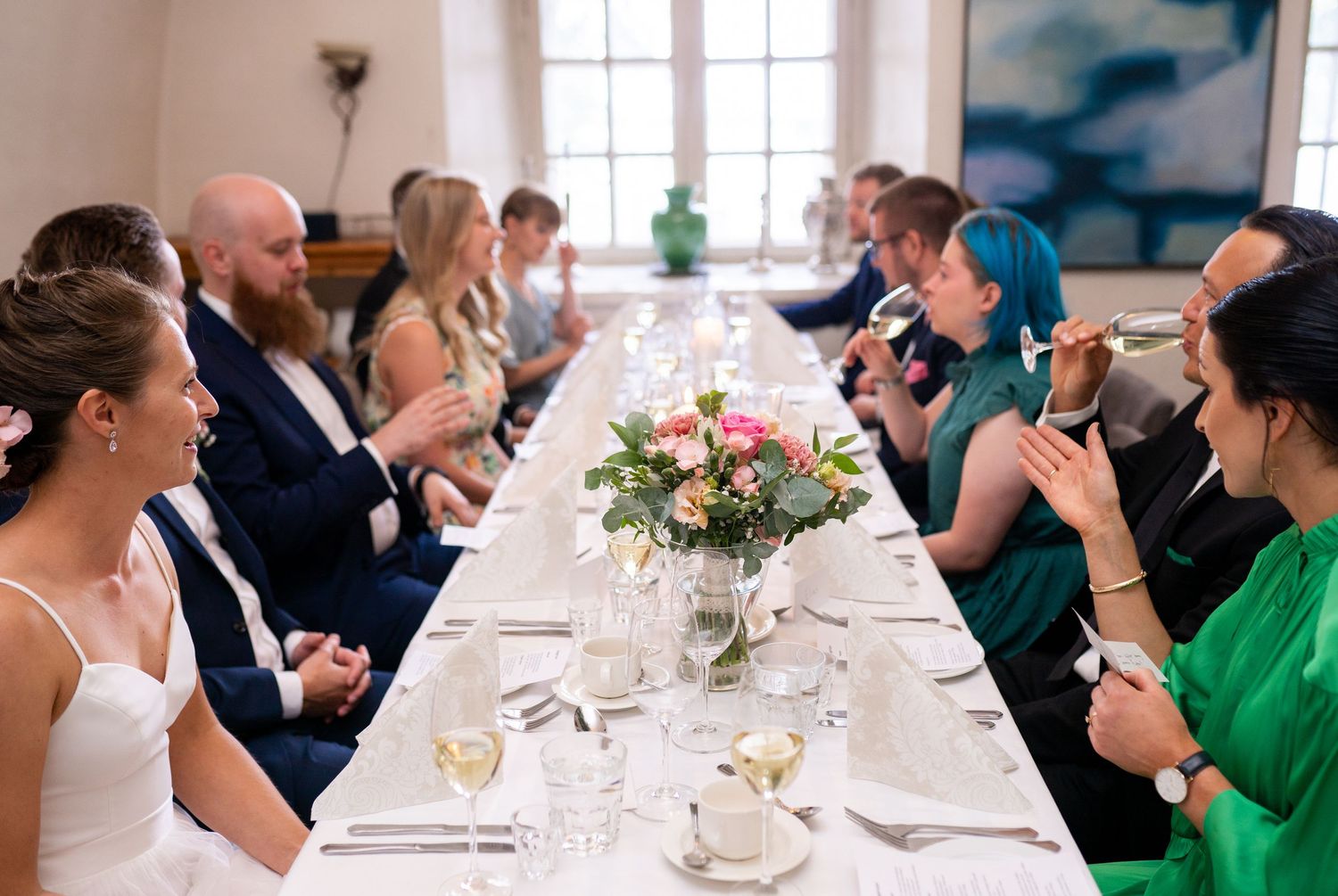 Wedding guests seated along a long white-clothed table with pink floral centerpiece during reception.