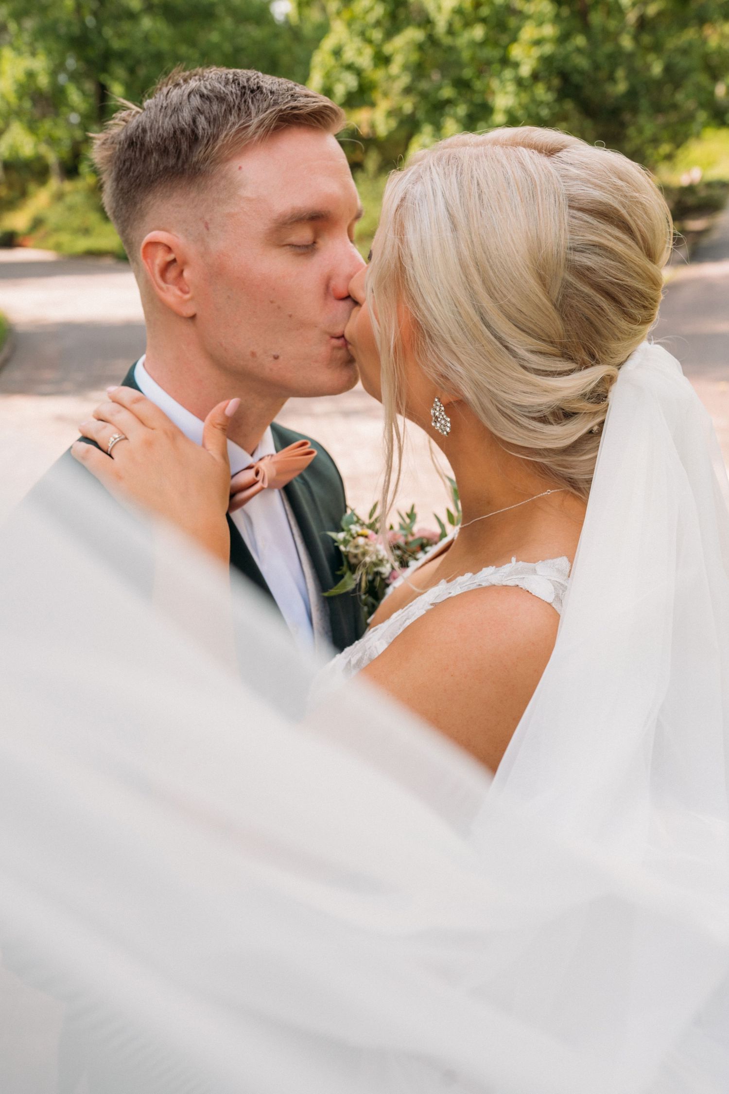 Bride and groom sharing romantic kiss outdoors with veil flowing in summer garden setting.
