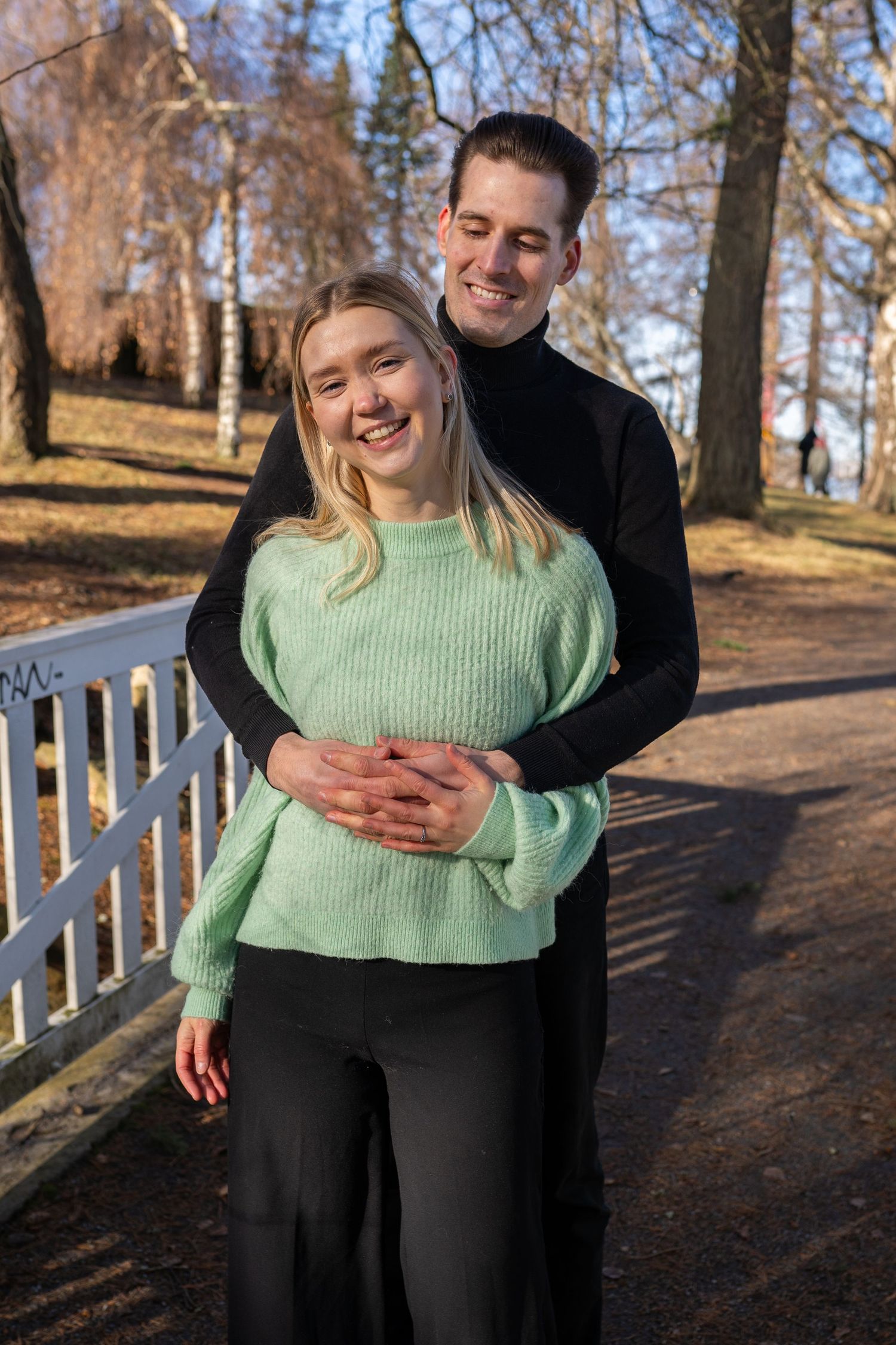 Happy couple embracing in a sunny park, woman in mint green sweater smiling, man in black turtleneck hugging her from behind.