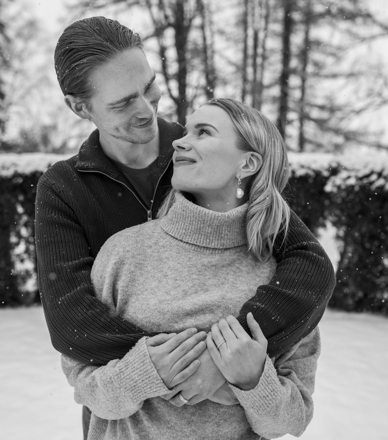 Black and white photo of a couple embracing and smiling in a snowy winter setting, wearing cozy sweaters.