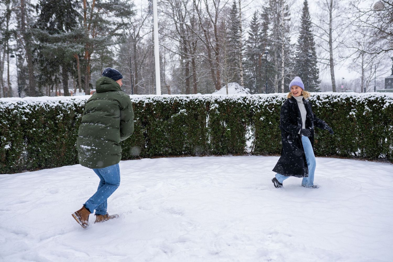 Two people playing in the snow in a winter park, wearing warm coats and hats amid snow-covered trees.
