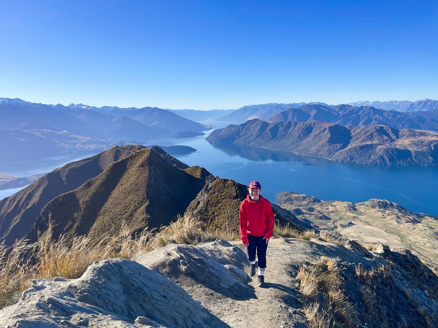 Woman in red jacket poses on rocky mountain trail with breathtaking lake and alpine scenery behind her.