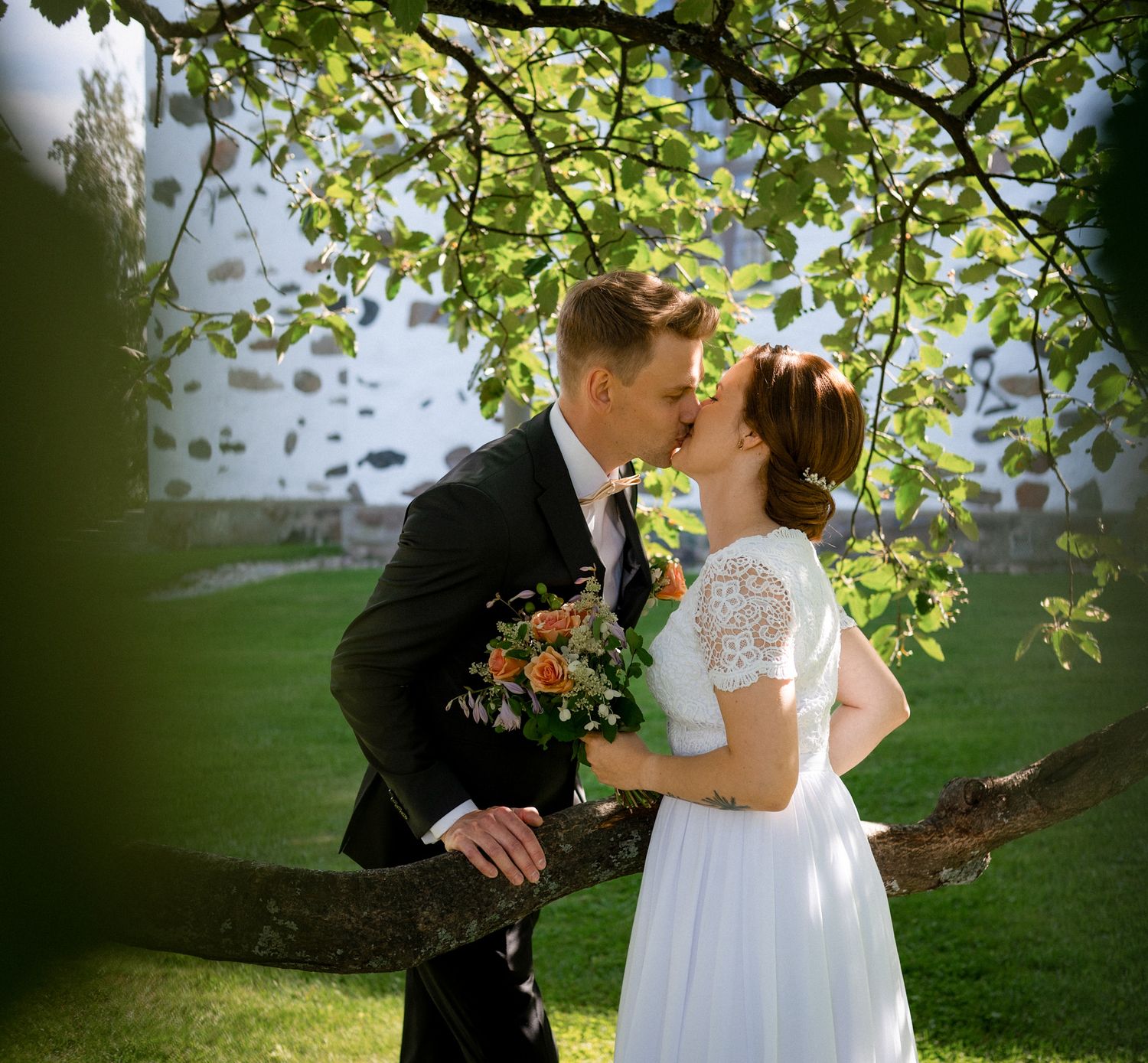 Bride and groom share a romantic kiss under green tree branches in a sunlit garden on their wedding day.
